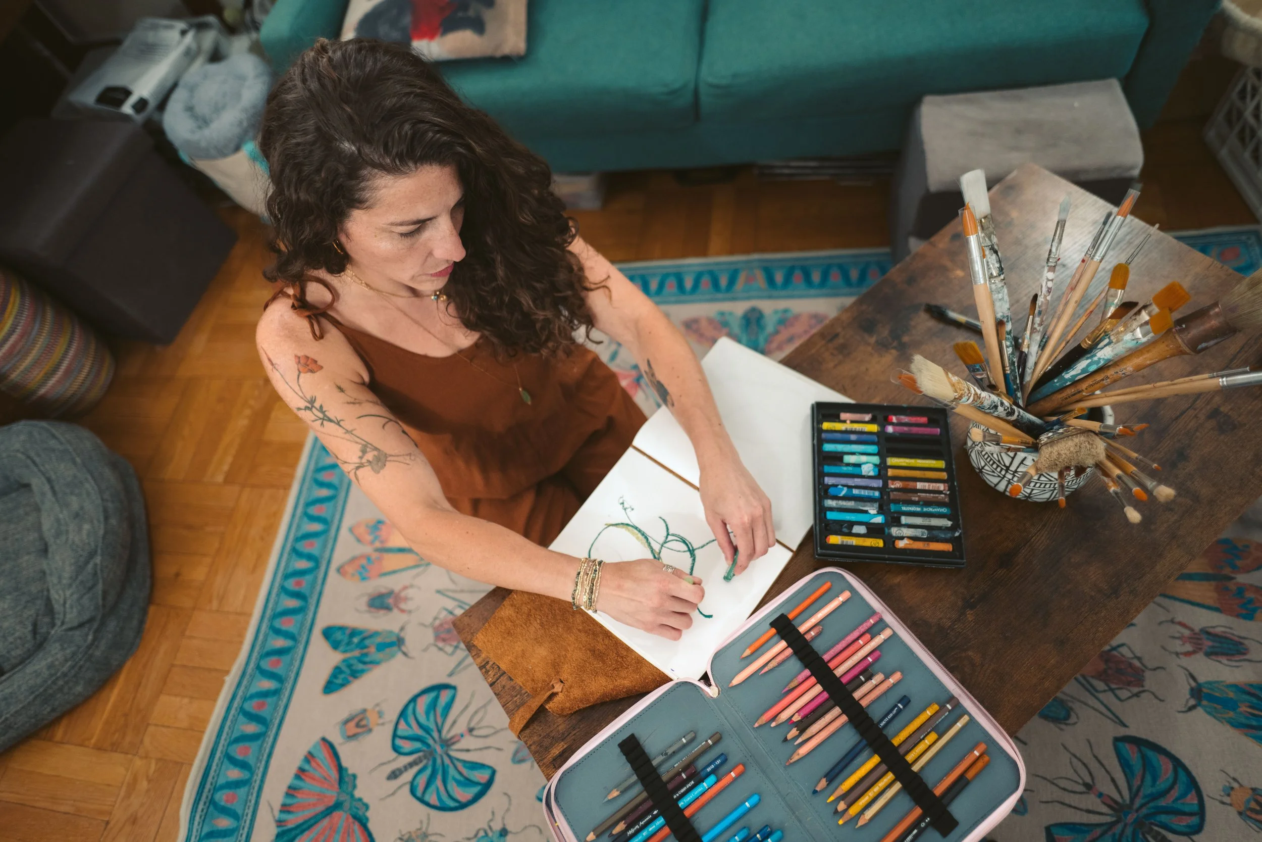 Abby Bramnick, Manhattan Art Therapist, sitting at a wooden table, drawing with colored pencils on a sketchbook, surrounded by art supplies including a container of paintbrushes, colored pastels, and a colored pencil case, with a teal couch.