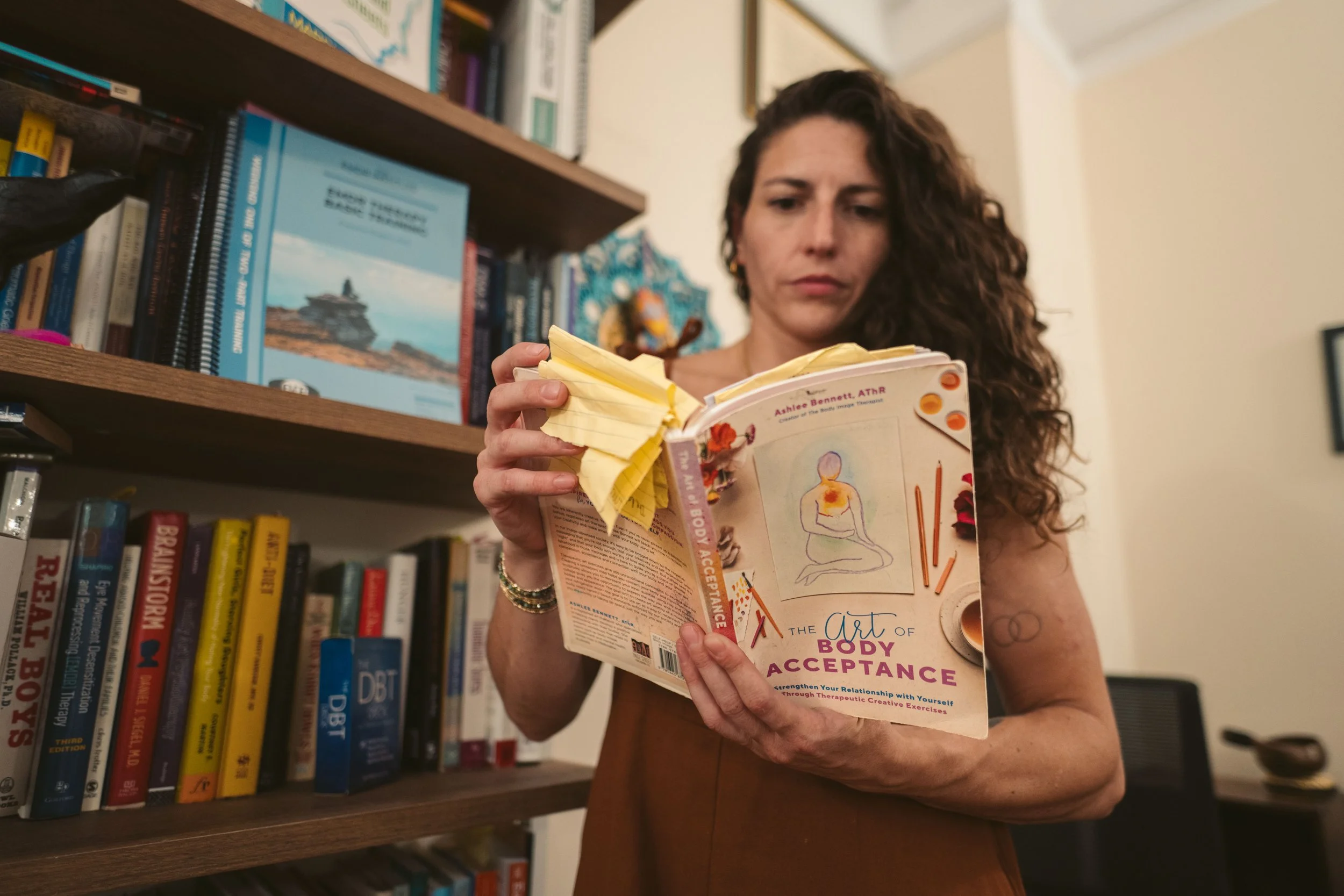Abby Bramnick, Manhattan Art Therapist and Eating Disorder Therapist, with curly brown hair is reading a book titled 'The Art of Body Acceptance' in a room with a bookshelf filled with various books.