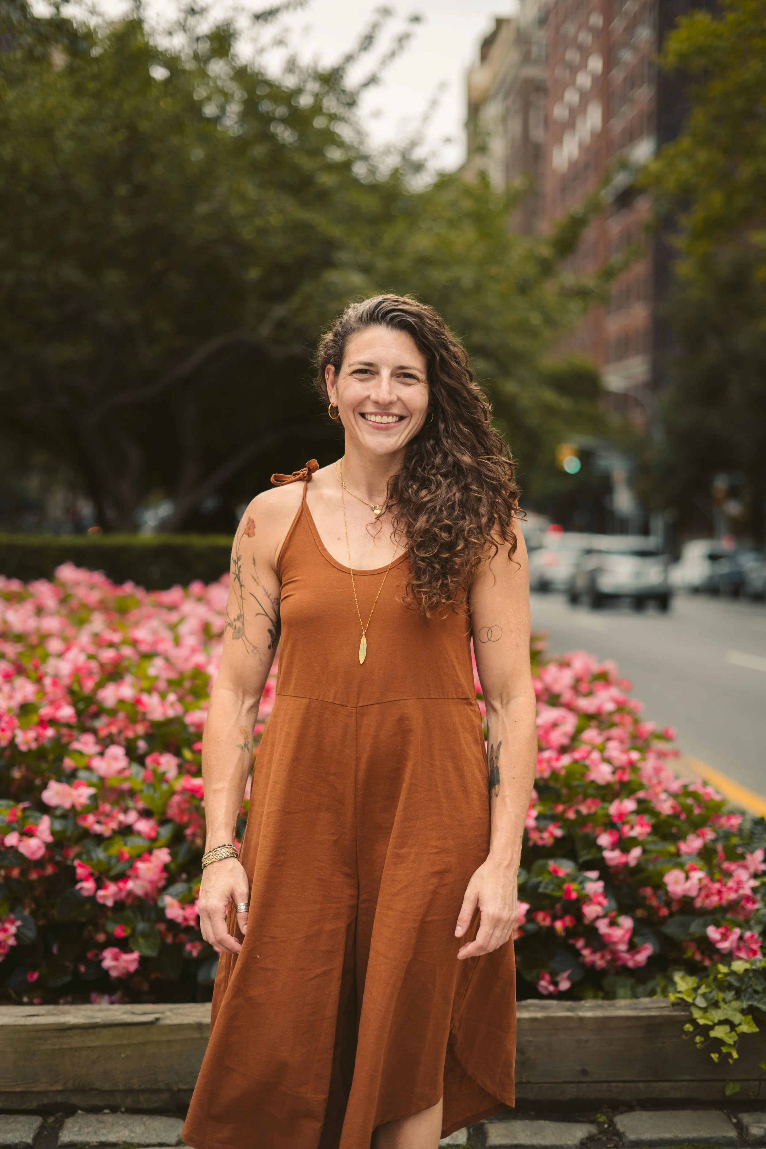 A woman with long, curly brown hair smiling while standing in front of pink flowers on a city sidewalk, wearing a brown dress and jewelry.