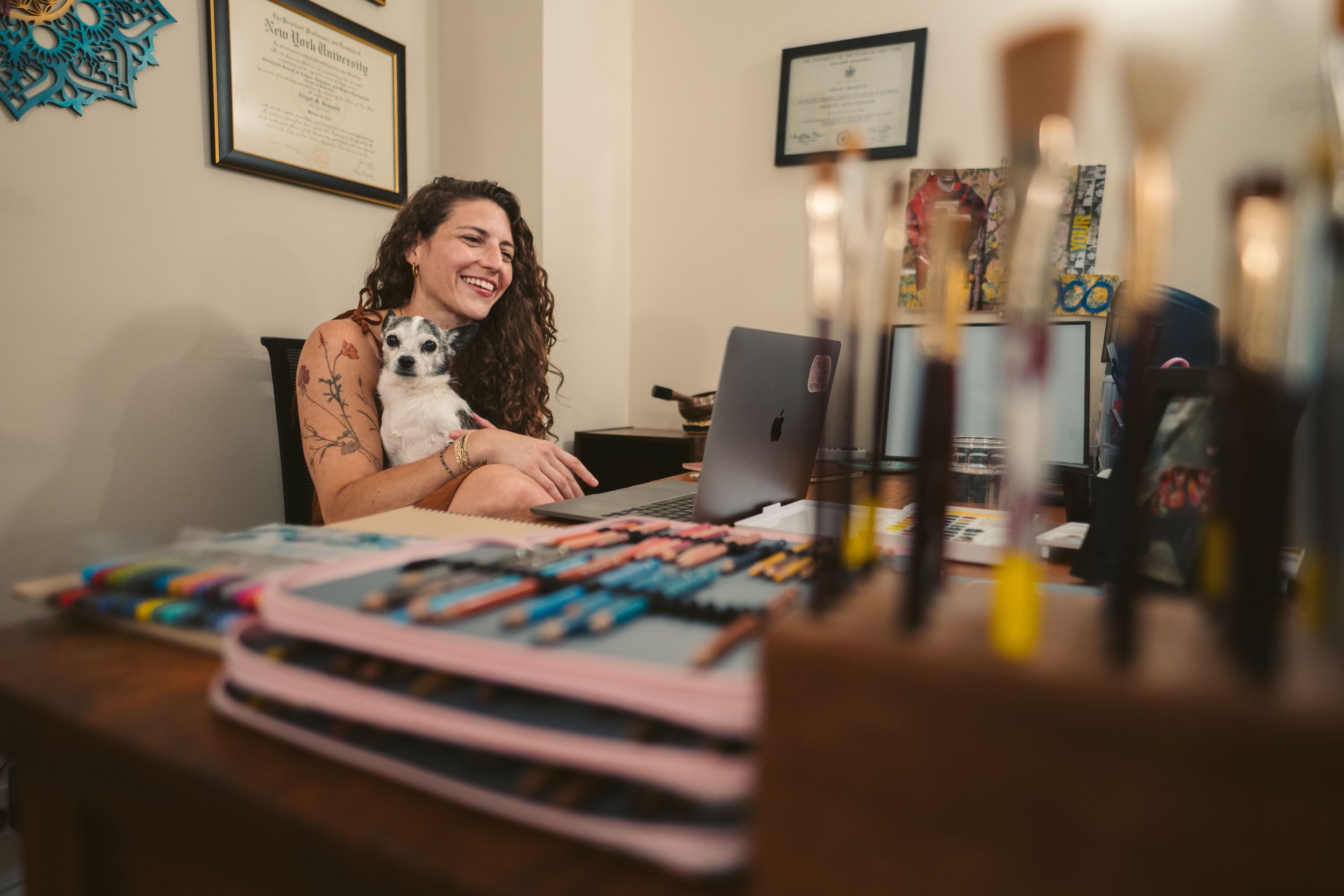 A woman with curly hair and tattoos on her arm is smiling while sitting at a desk and holding a small black and white dog. On the desk are various colored markers, notebooks, and a laptop. Behind her are framed diplomas on the wall and some artwork.