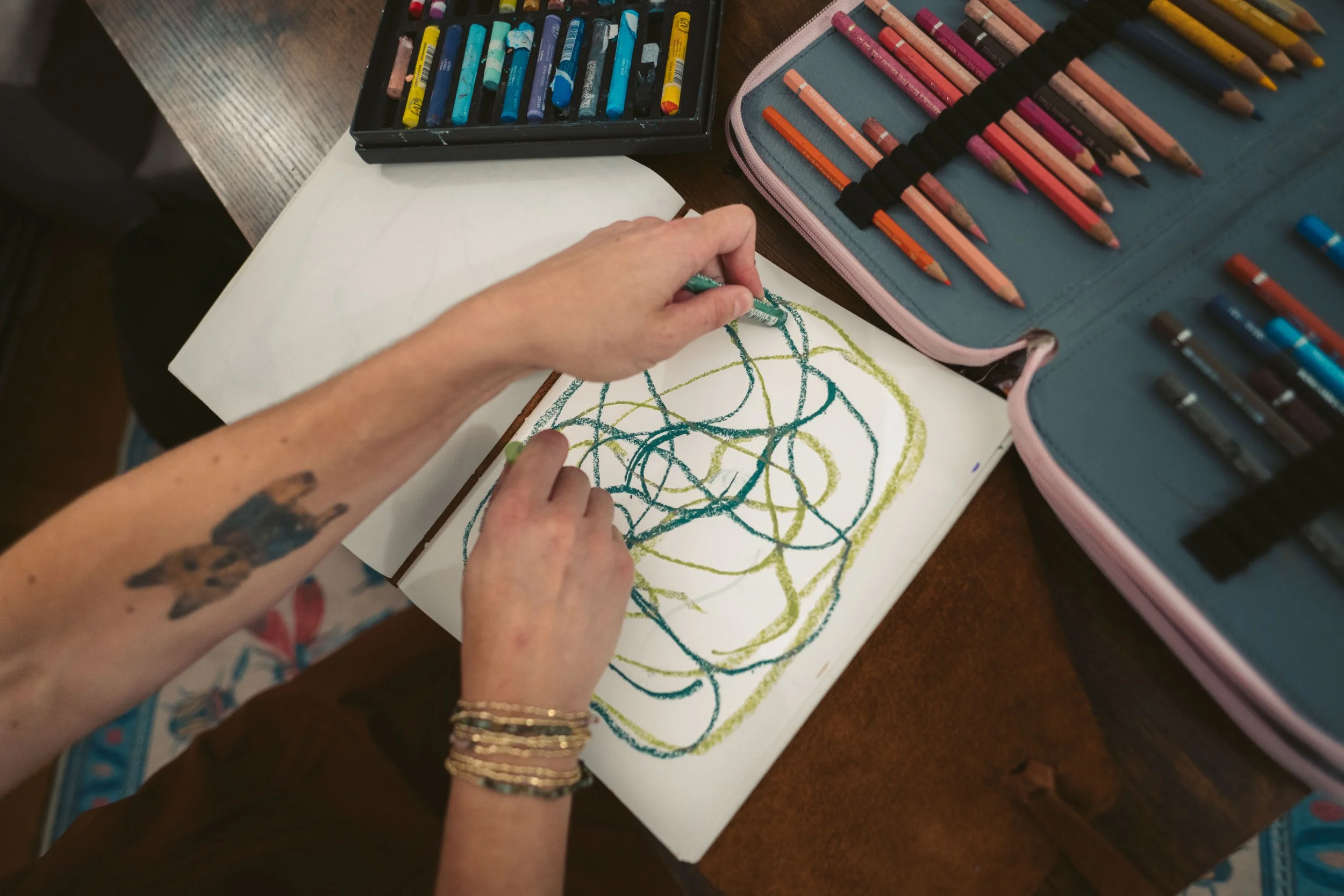 Abby Bramnick, Manhattan Art Therapist, drawing colorful scribbles on paper with crayons and pencils, surrounded by open colored pencil cases on a wooden table.