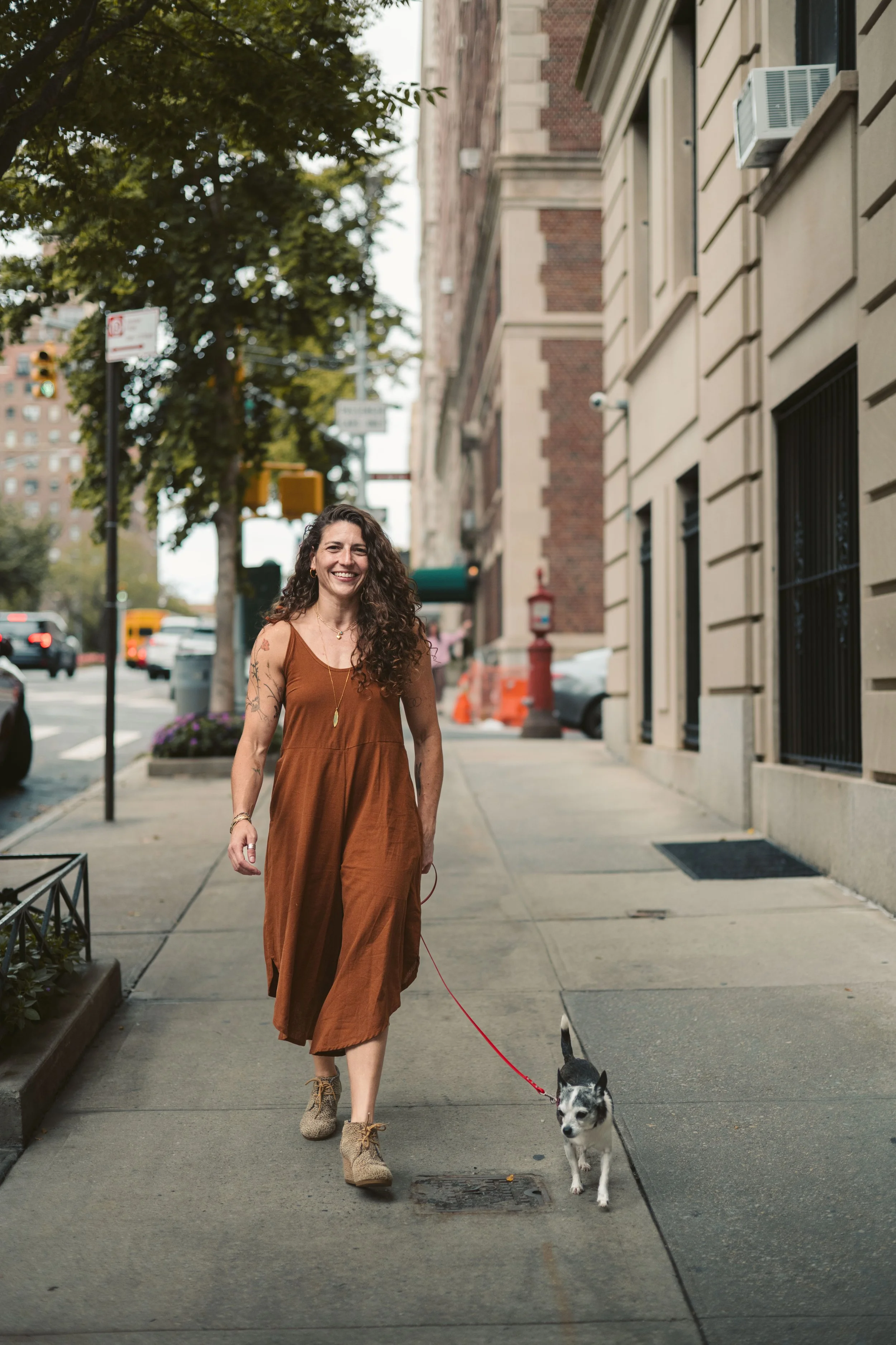 Abby Bramnick, Manhattan Art Therapist and Eating Disorder Therapist, walking a small black and white dog on a city sidewalk, smiling. Buildings and trees in the background.