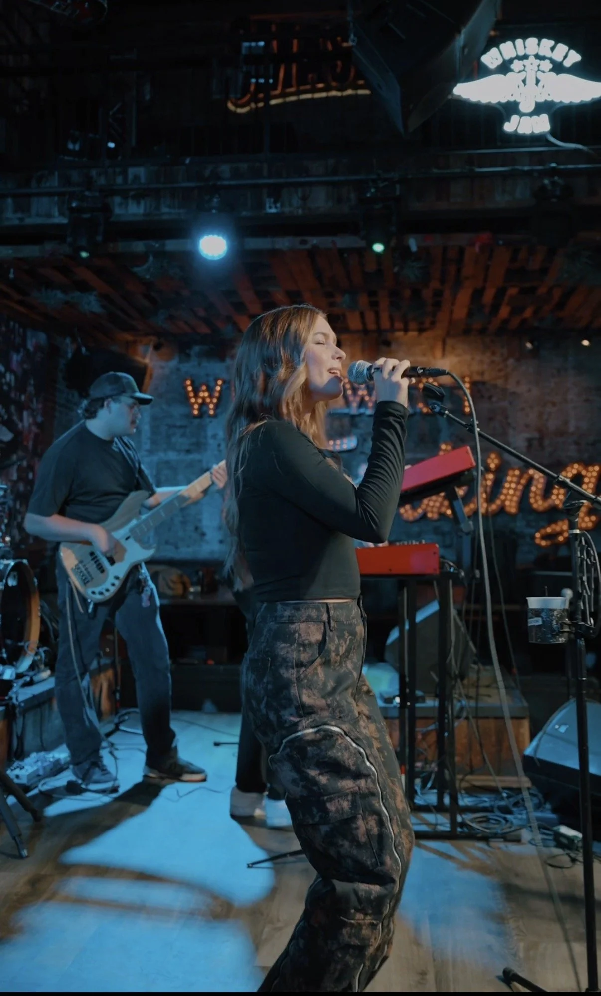 A female singer with long hair performing on stage with a microphone, accompanied by a bassist in the background, in a dimly lit venue with brick walls and decorative lighting.
