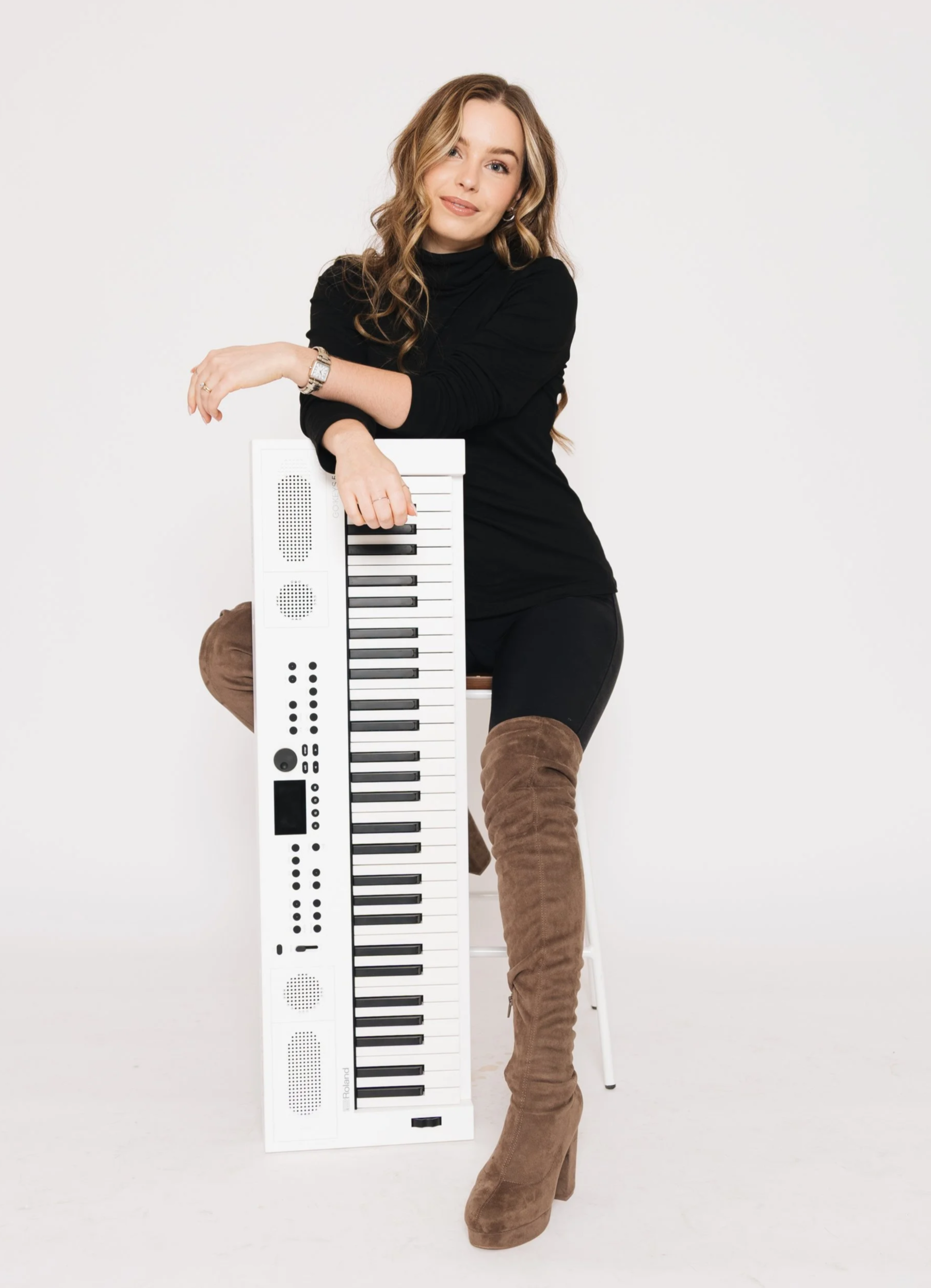 A woman with long, wavy, blonde hair dressed in a black top and black pants, sitting on a white chair, resting her arms on a white electronic keyboard with black and white keys, against a plain white background, wearing brown over-the-knee boots.