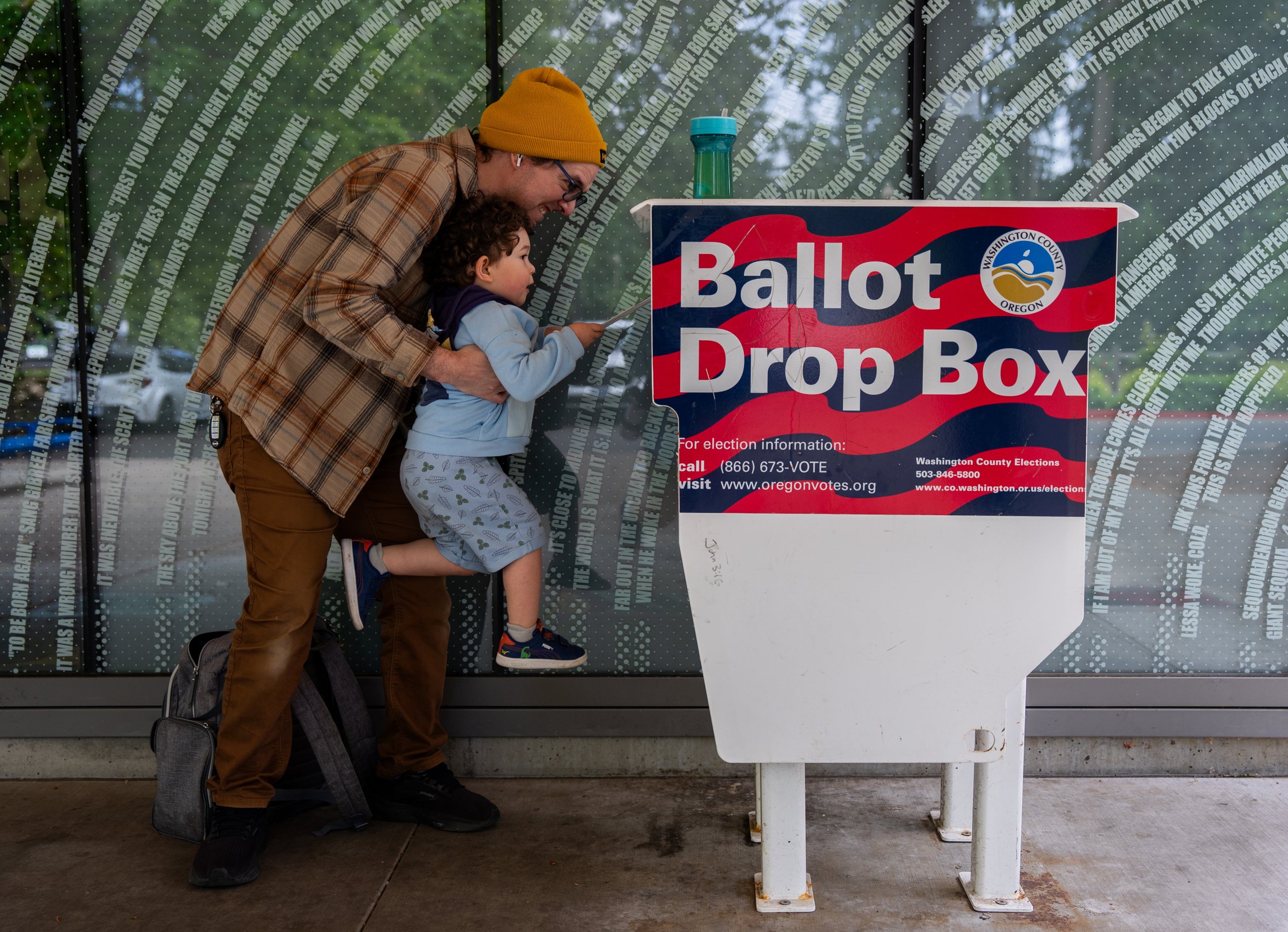Matt lifts up his three-year-old son, Theo, to cast his ballot into the Hillsboro Public Library-Shute Park ballot drop box in May 2026.  Eli Imadali / OPB