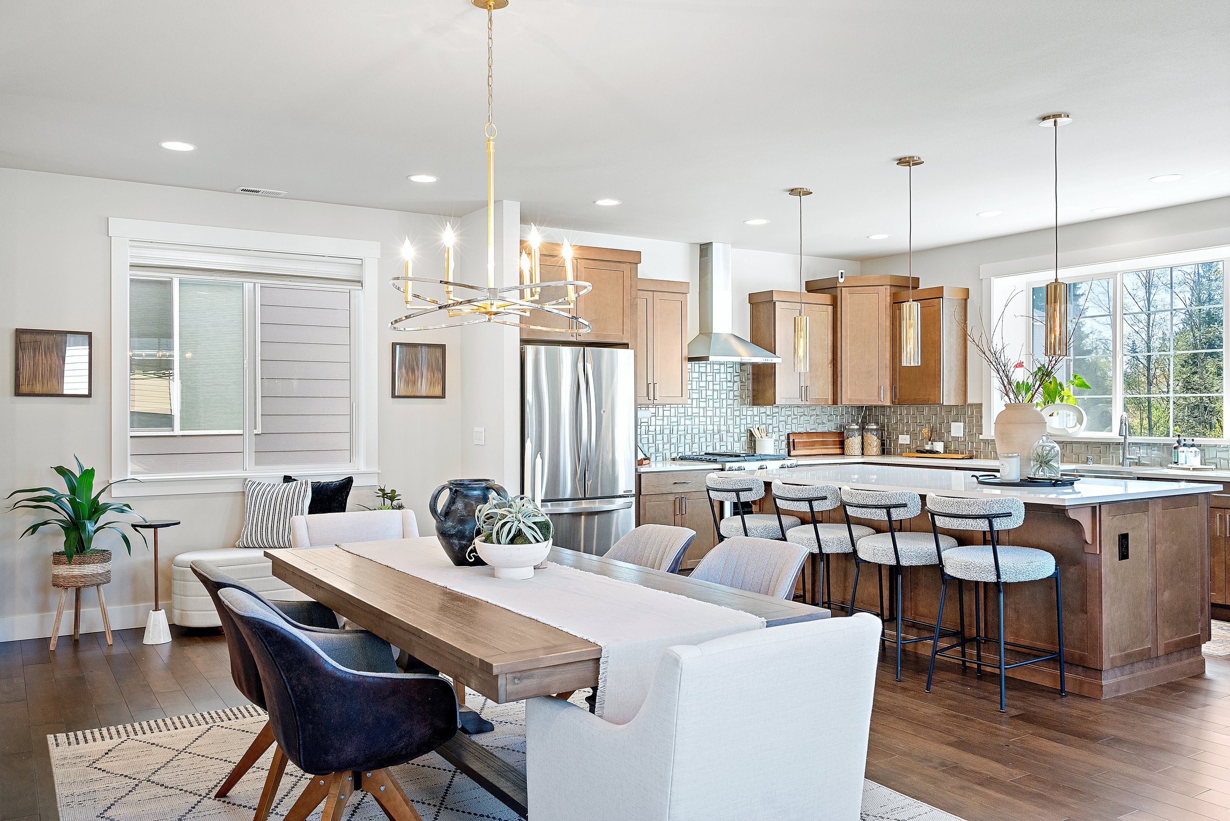 Modern kitchen and dining area with wooden cabinetry, stainless steel appliances, island with bar stools, large dining table, pendant lighting, and decorative plants.