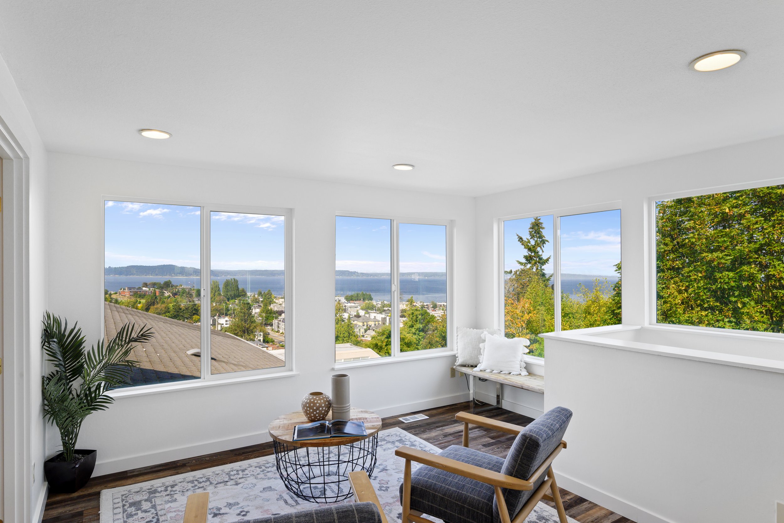 Bright room with large windows offering a scenic view of trees and a water body. Furnished with a couple of chairs, a small round table, and a bench with cushions. The room has wooden flooring and a potted plant in the corner.