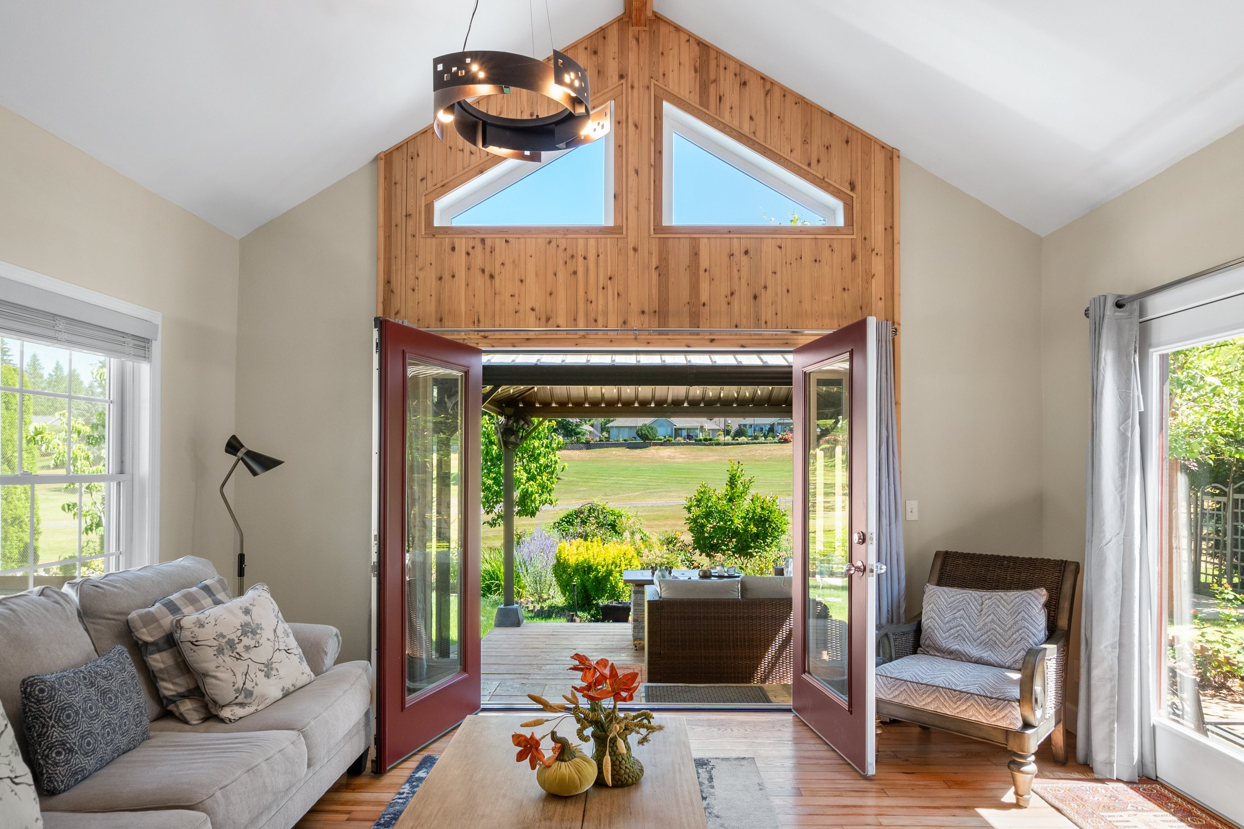 Cozy living room with gray sofa, patterned cushions, modern floor lamp, and wooden coffee table. Open French doors lead to a patio with outdoor furniture and a scenic view of a garden and lawn. Contemporary chandelier and natural light enhance the sp