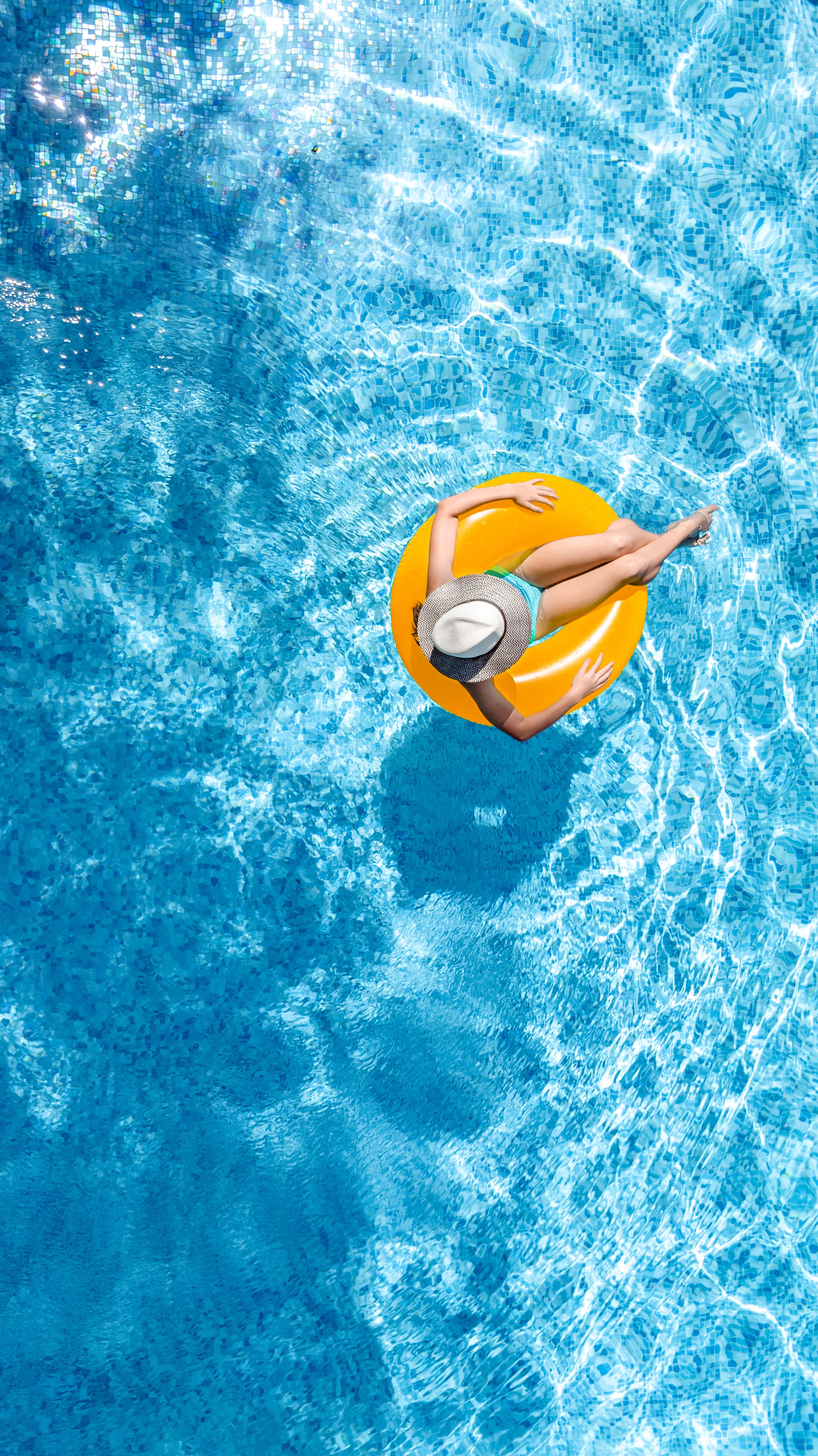 A woman in a swimsuit and wide-brimmed hat relaxing on an orange inflatable ring in a swimming pool with mosaic tile bottom.