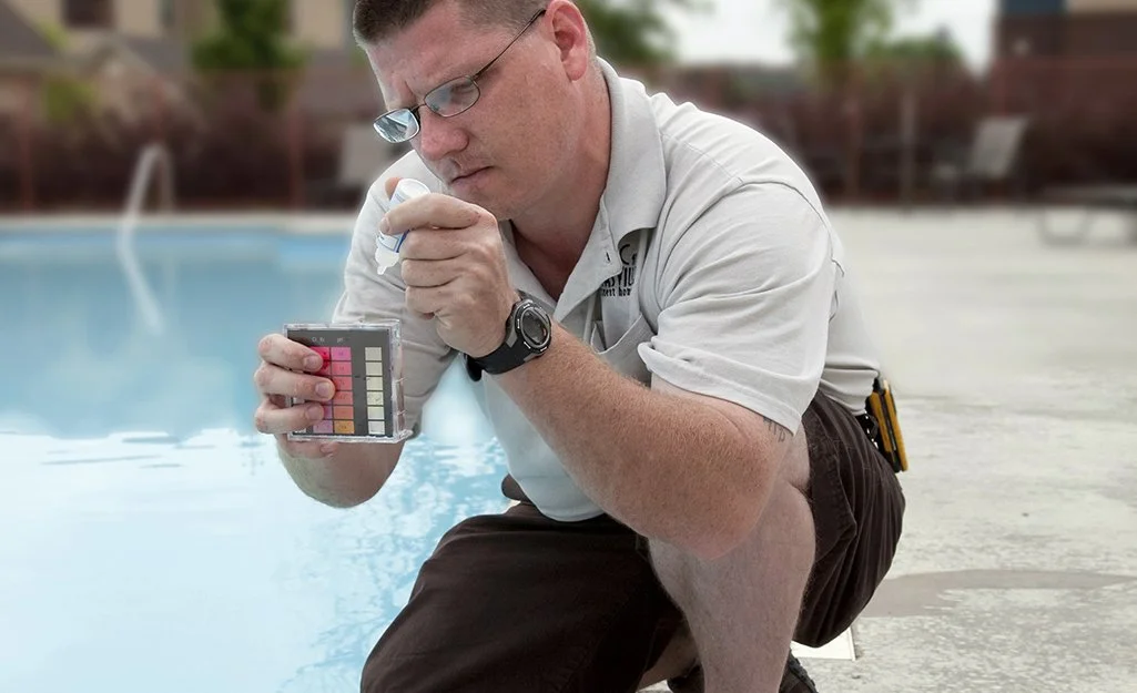 A man kneeling by a swimming pool, examining a pH test strip using a color comparison chart.