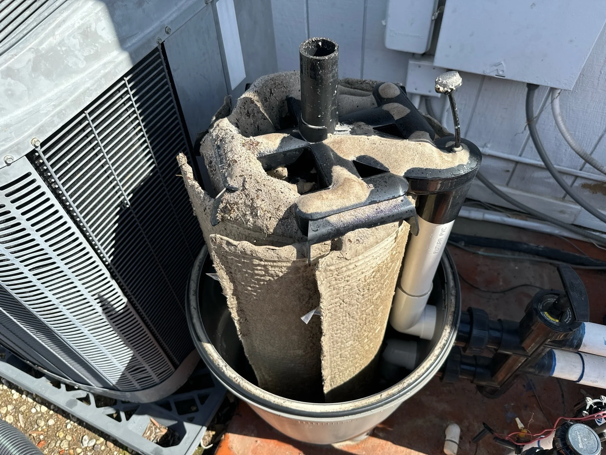 An outdoor scene showing a damaged air conditioning unit, a bucket with concrete and debris, a piece of black pipe, and a shock absorber on a brick surface.