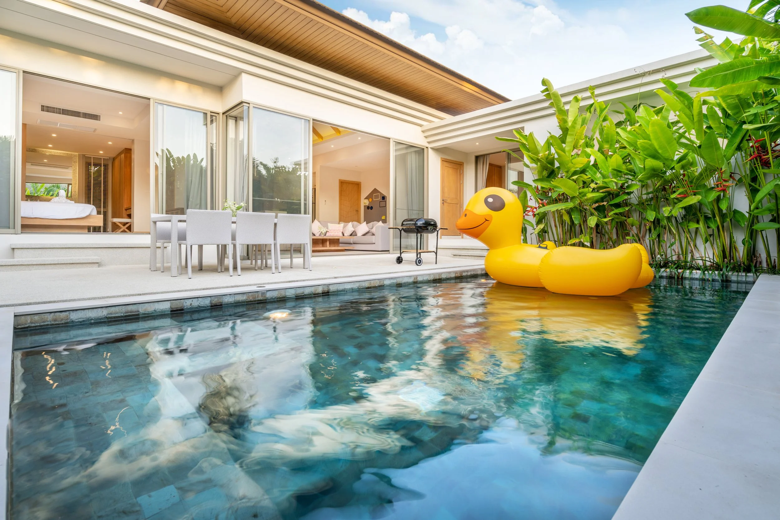 A modern house with large glass sliding doors opening to a patio with a dining area. In the pool, there's a yellow inflatable duck float. The house has a wooden roof overhang, and there are lush green plants on one side. The interior of the house is visible through the glass doors, showing a bedroom and a living room.