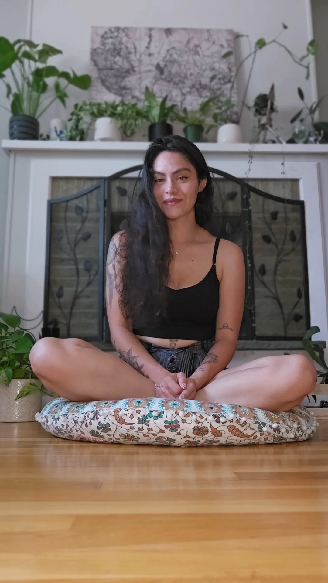 A woman with long black hair, sitting cross-legged on a patterned cushion in front of a fireplace decorated with plants. She is wearing a black crop top and striped shorts, with visible tattoos on her arms.