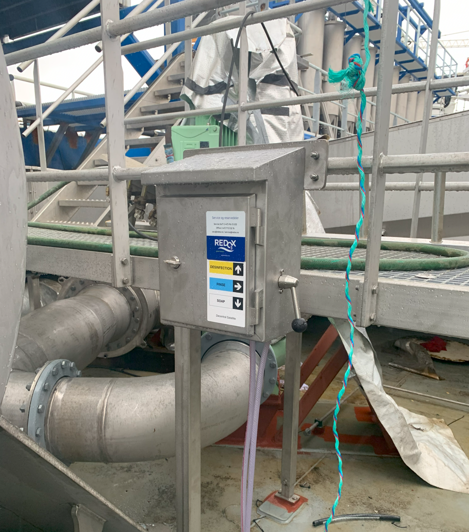Industrial setup with a control box labeled 'REDCOX' mounted on a metal frame next to large pipes, with labeled buttons for disinfection, rinse, and soap. A twisted blue and turquoise rope hangs beside the setup.
