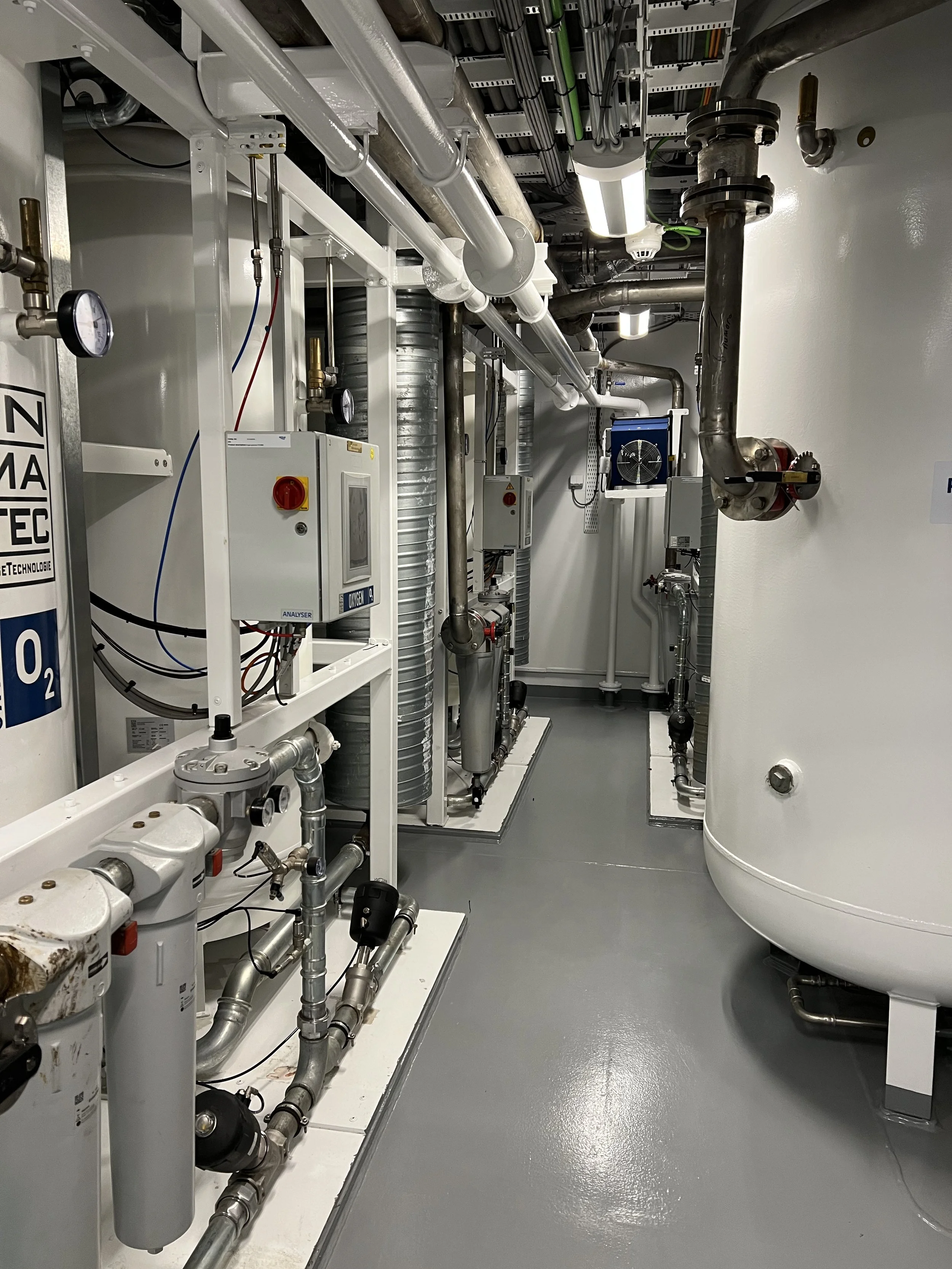 A mechanical room with various pipes, valves, gauges, and equipment, including large vertical tanks and control panels on a gray floor.