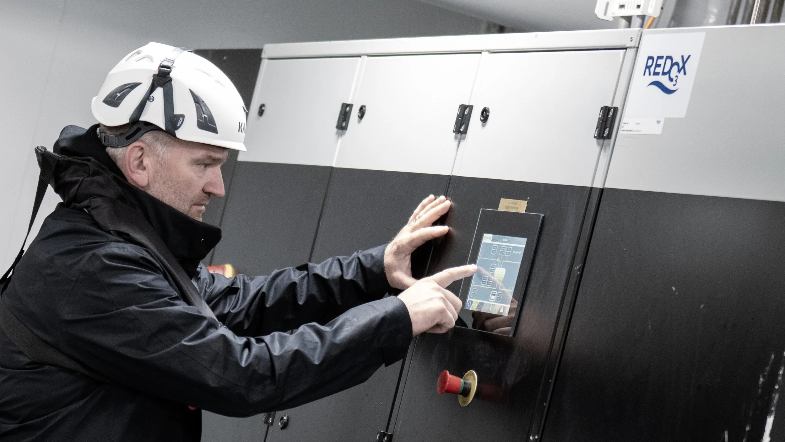 A man wearing a safety helmet and black jacket operating a control panel in an industrial setting.