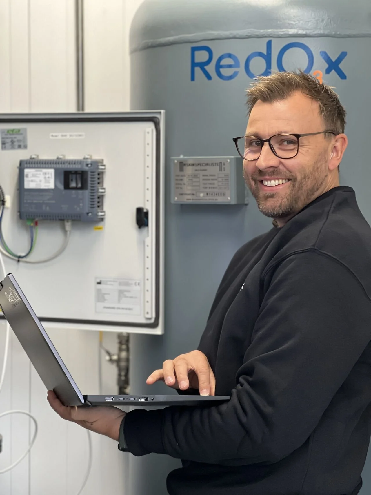 A man with glasses and a beard working on a laptop in front of industrial electrical equipment, with a large blue water tank labeled 'Redox' in the background.