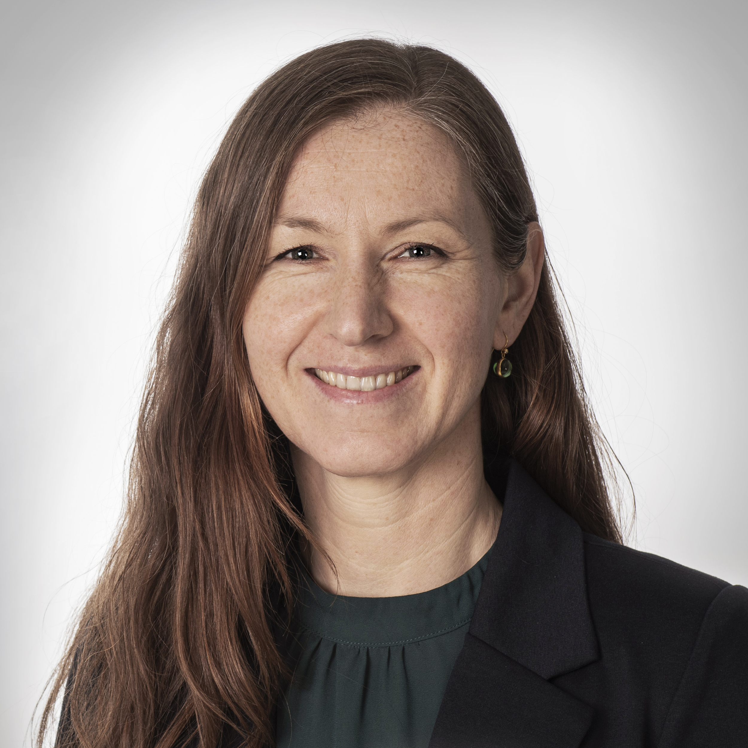Portrett av en smilende kvinne med lang, brunt hår, iført svart blazer og mørkegrønn topp, mot en lys bakgrunn.