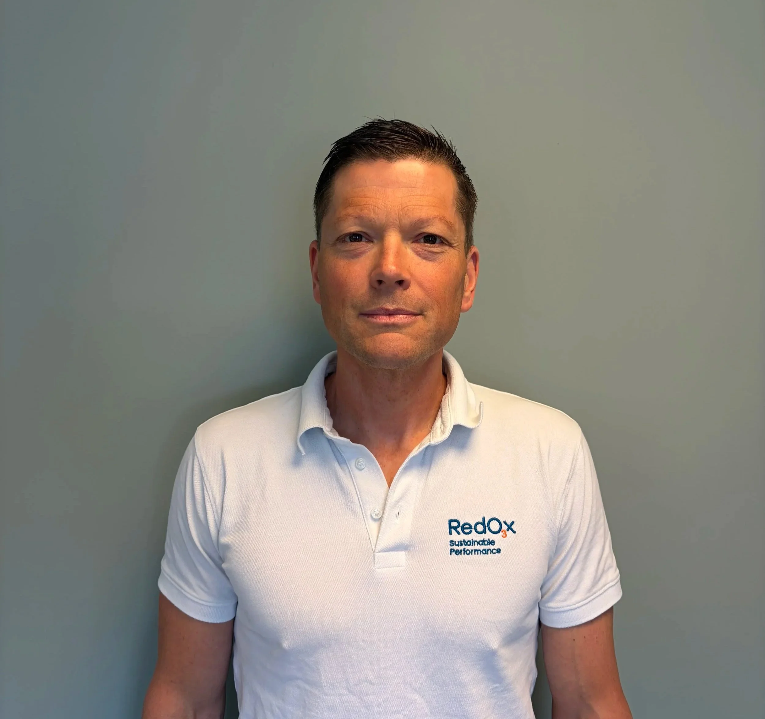 A man taking a selfie indoors, wearing a black polo shirt with 'Redox' logo, with a landscape painting and a potted plant on a cabinet in the background.