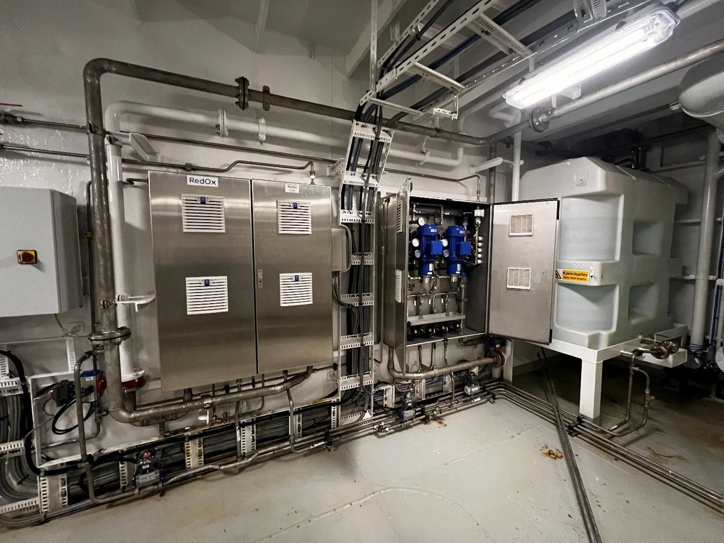 Tekniske installasjoner med kontrollskap, pumper og rør i en industribygning.