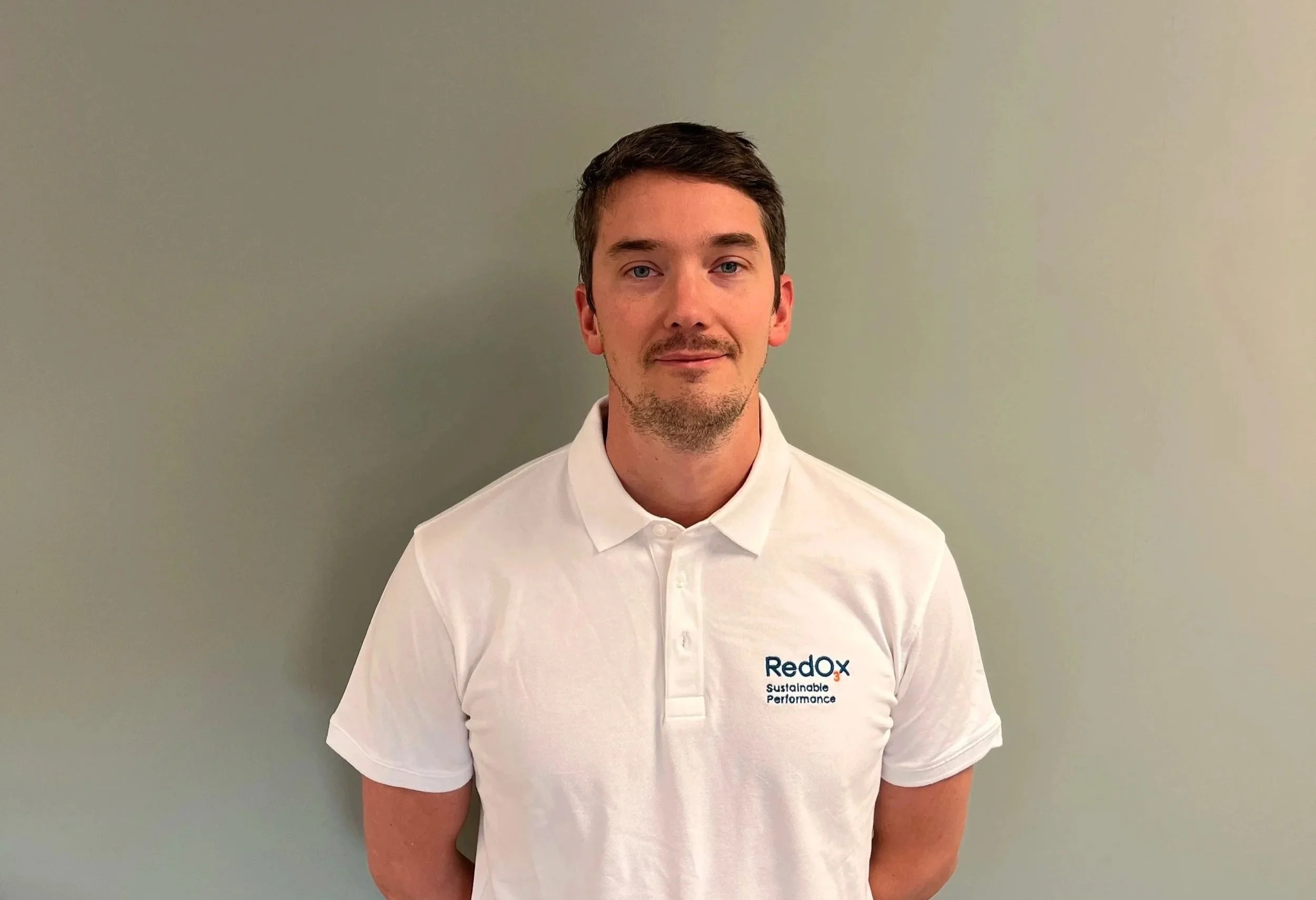 A young man with short brown hair and a light beard smiling, wearing a blue collared shirt, outdoors near a body of water with blurred buildings in the background.