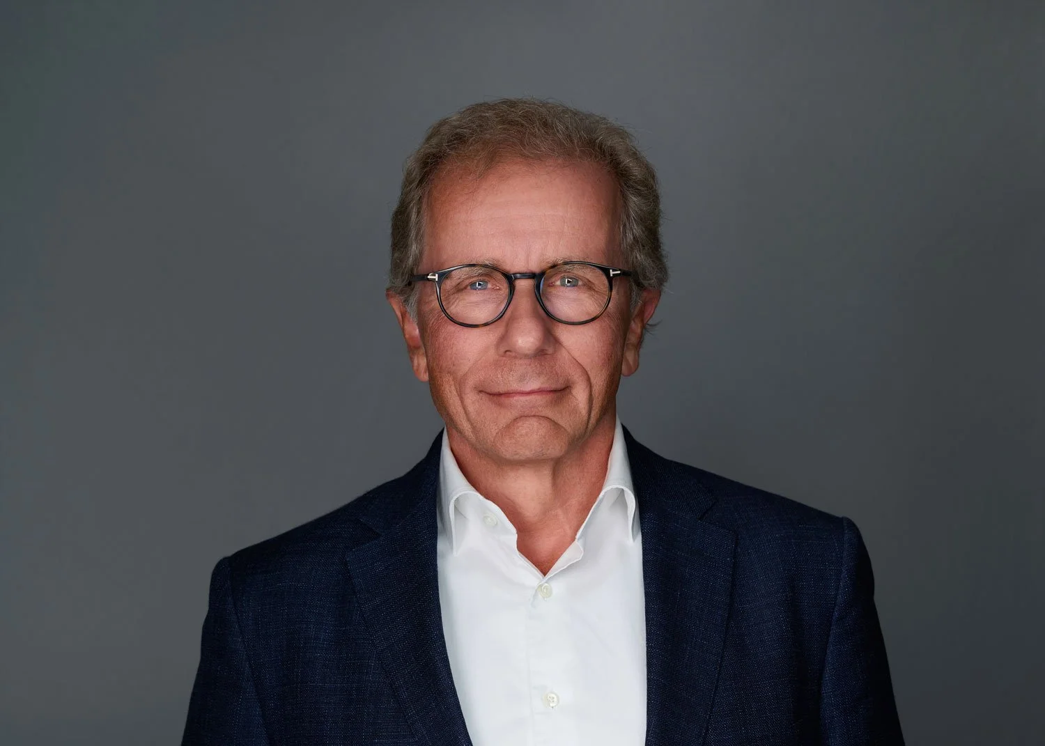 Headshot of a middle-aged man with glasses, gray hair, wearing a dark blazer and white shirt, against a gray background.