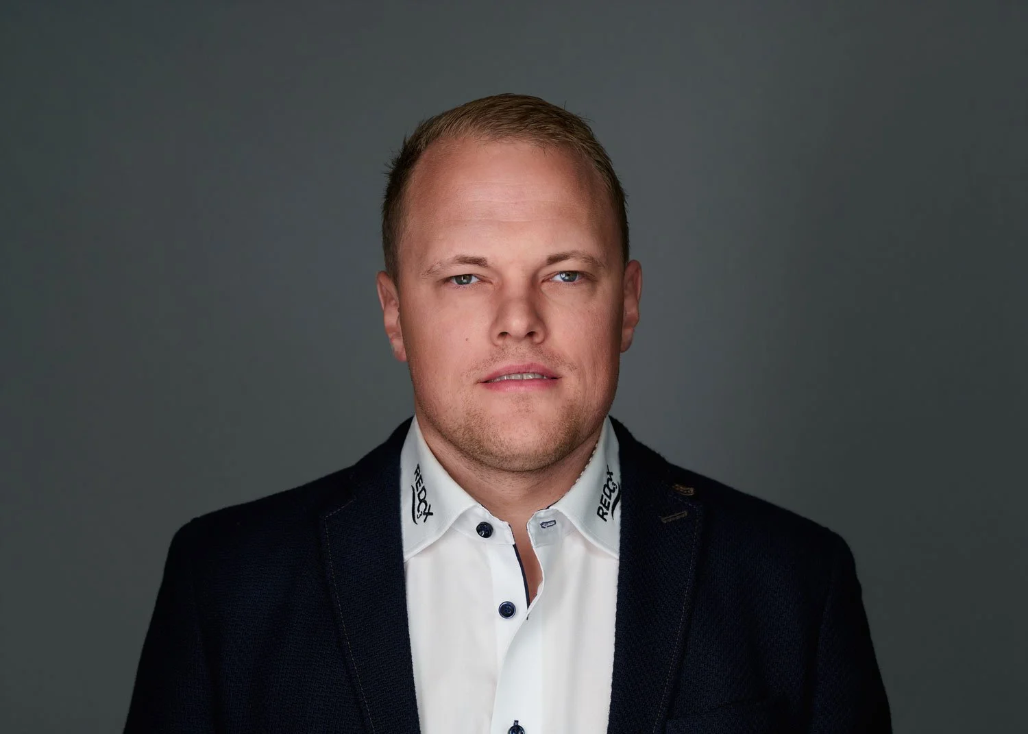 Portrait of a man with short blonde hair, blue eyes, wearing a dark suit and a white shirt with a high collar, against a gray background.