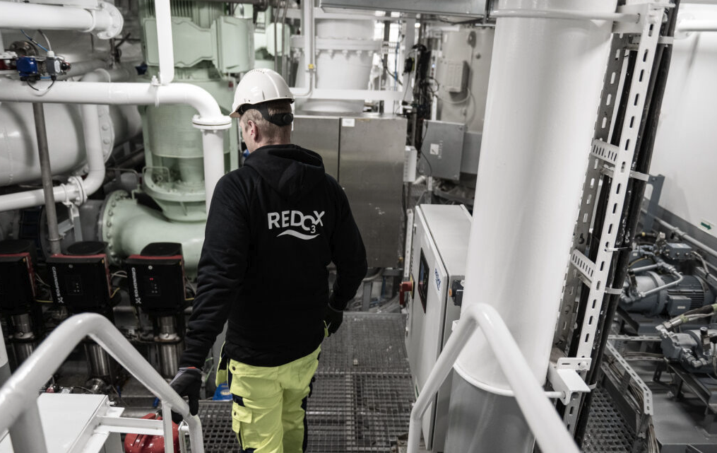 A worker wearing a white safety helmet and a black hoodie with 'REDCX3' on the back, in an industrial plant with pipes, tanks, and machinery.