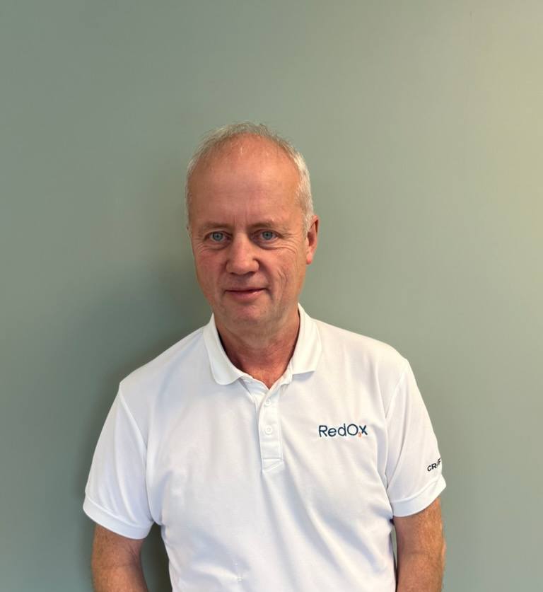 Portrait of a bald man with blue eyes wearing a light blue zip-up polo shirt, against a plain light gray background.