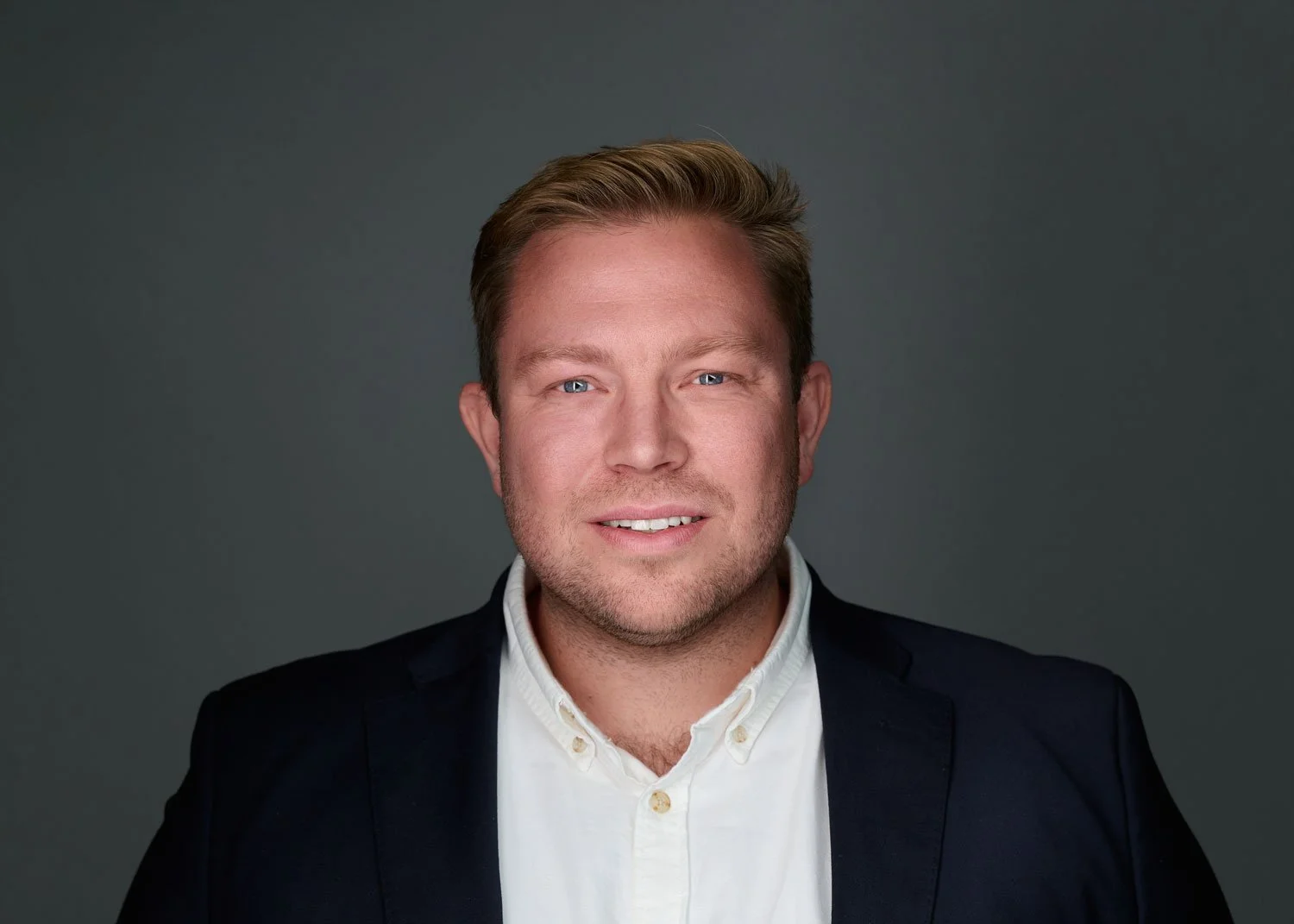 Headshot of a smiling man with blond hair, blue eyes, wearing a white shirt and dark blazer, against a gray background.