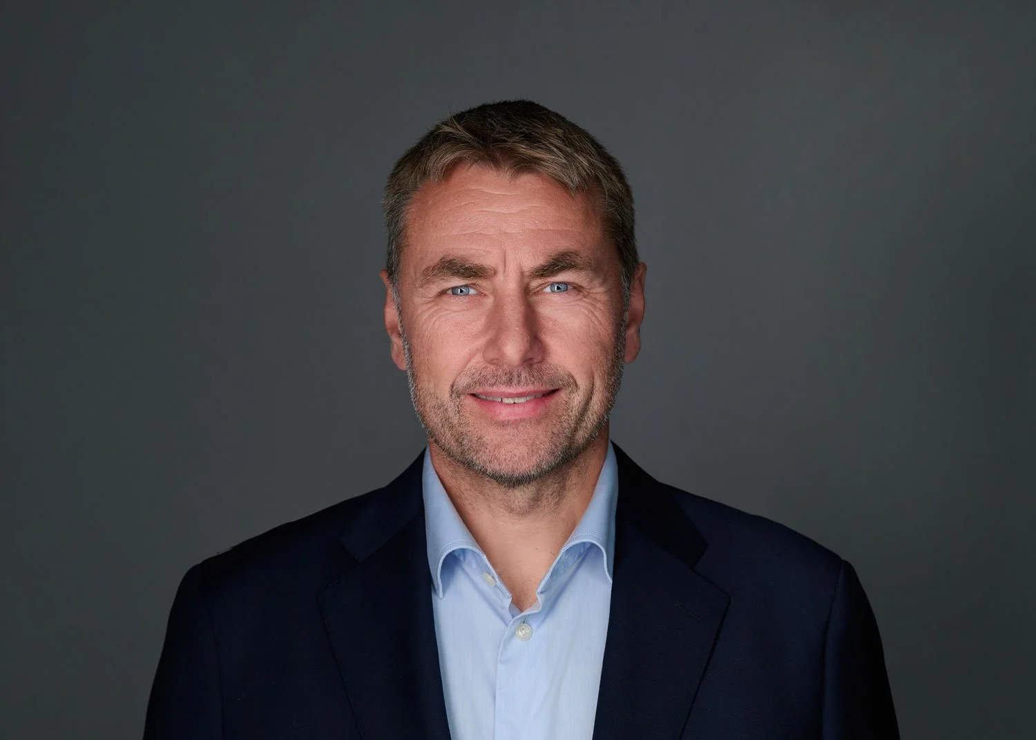 A middle-aged man with light brown hair, blue eyes, and a beard, wearing a dark suit jacket and light blue dress shirt, smiling lightly against a gray background.