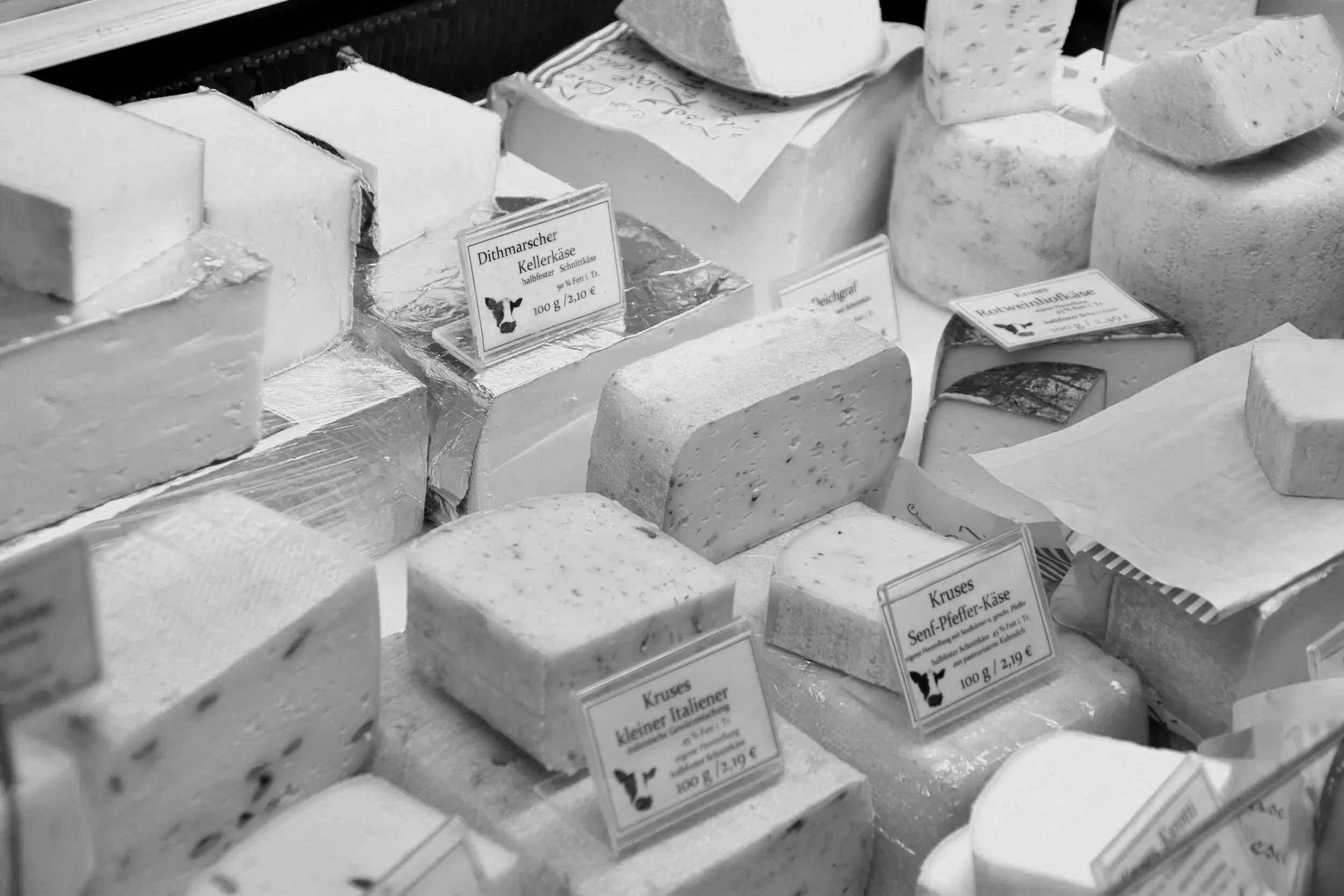 En utstilling av forskjellige typer ost, inkludert blåmuggost og urtekrydrede oster, med prisskilt som viser vekt og pris i euro.