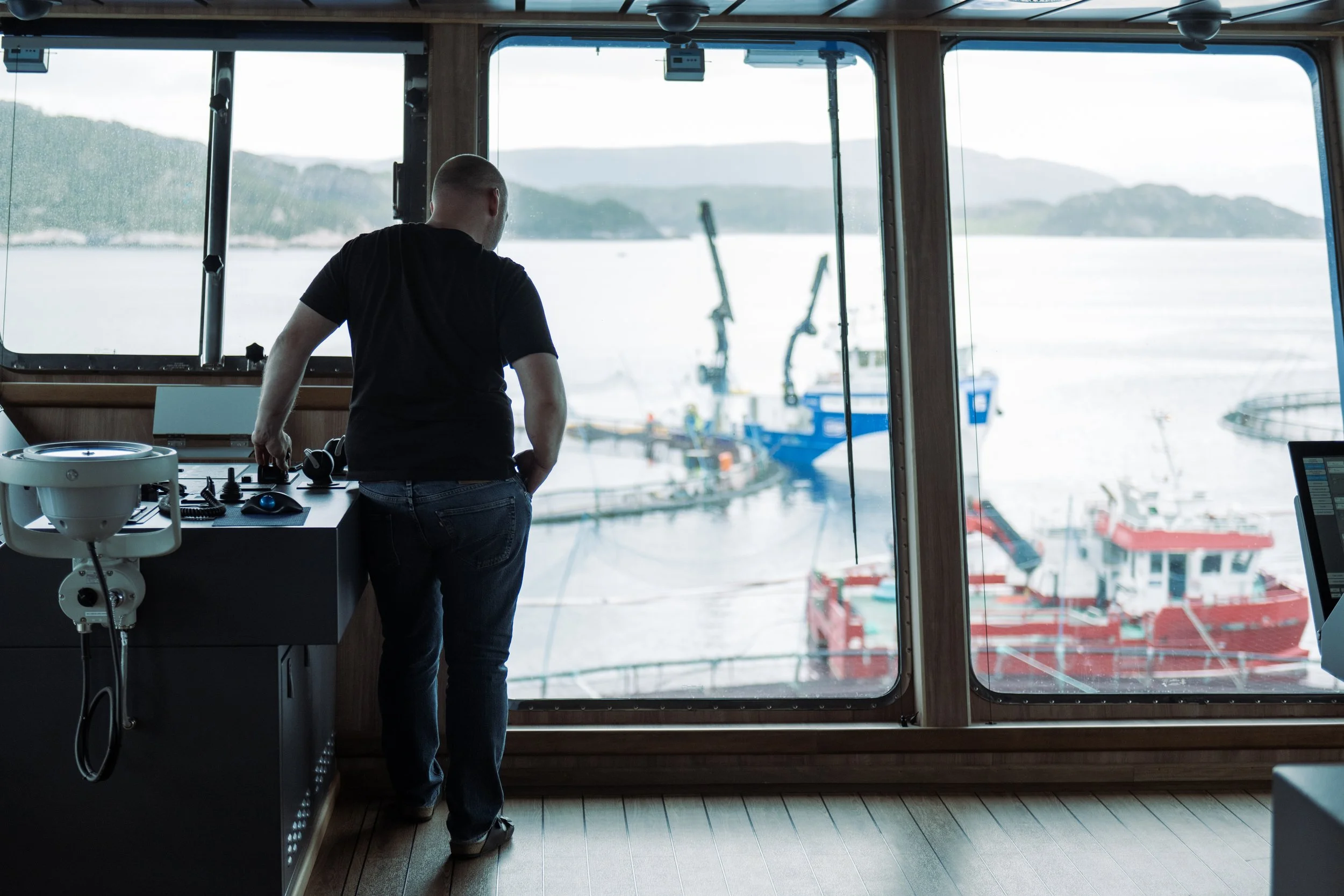 A man in a black shirt stands inside a boat's control room, looking out the large windows over a marina filled with boats.