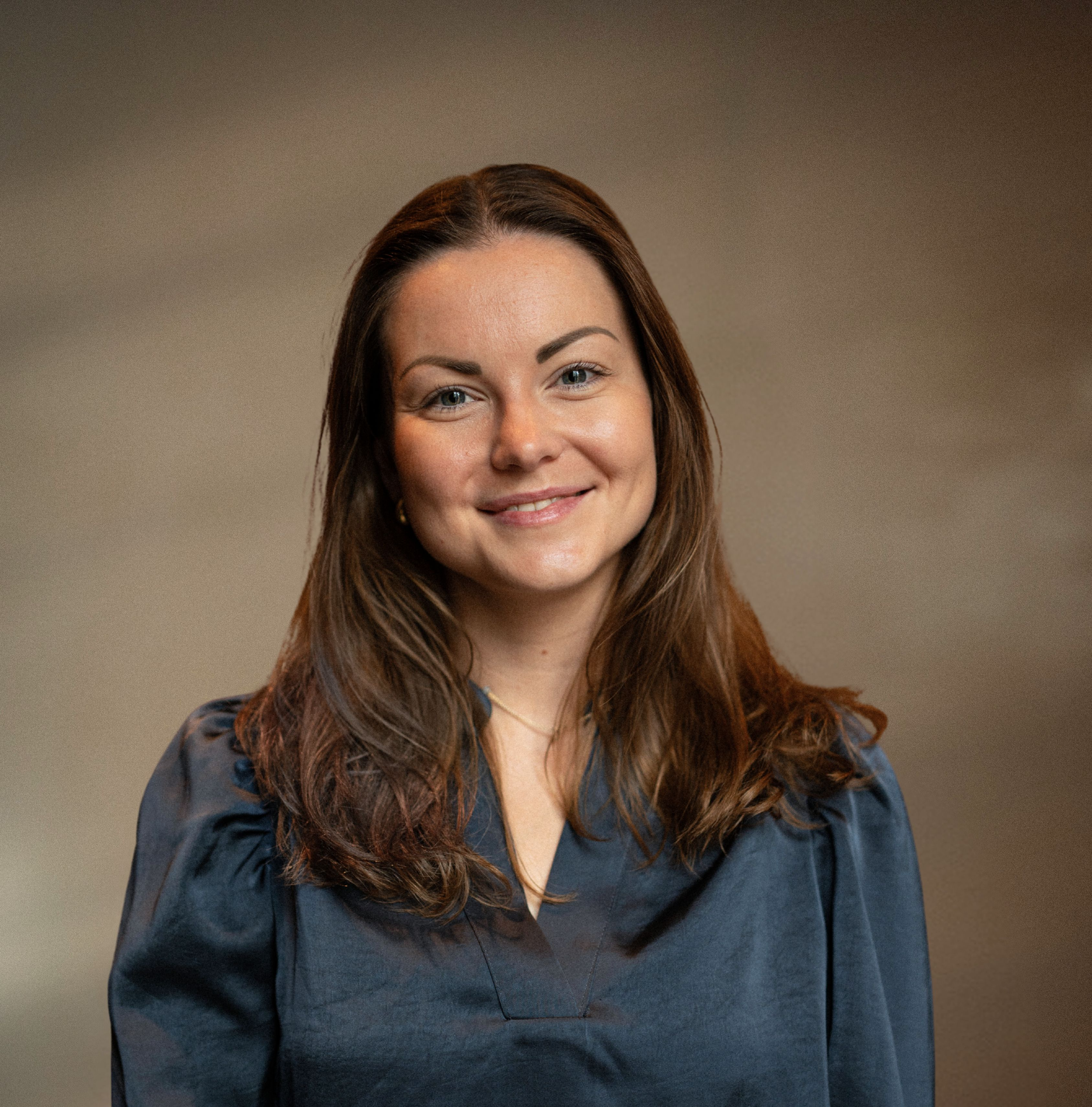 Portrett av en smilende kvinne med brunt hår, iført mørk blå kjole, mot en enkel, brun bakgrunn.