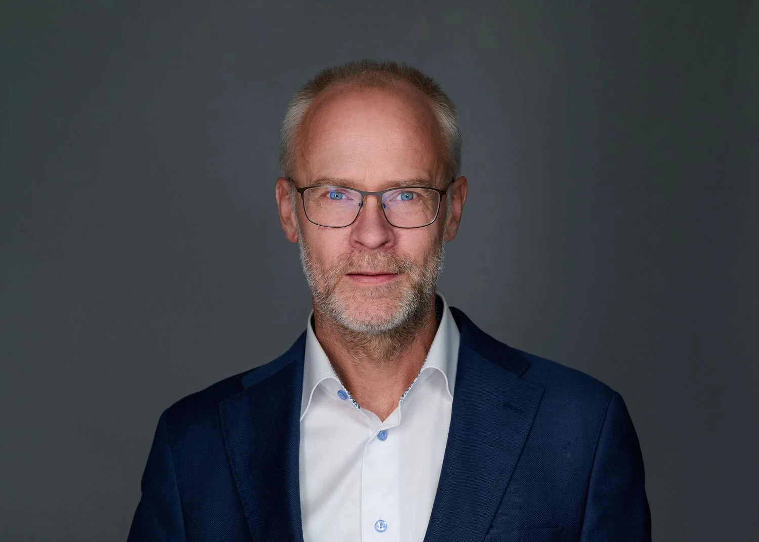 A professional man with glasses, gray hair, and a beard, wearing a dark blue suit and white shirt, posing against a gray background.