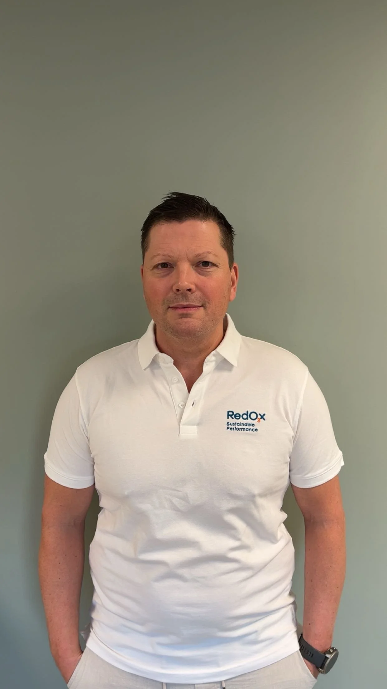 A man taking a selfie indoors, wearing a black polo shirt with 'Redox' logo, with a landscape painting and a potted plant on a cabinet in the background.