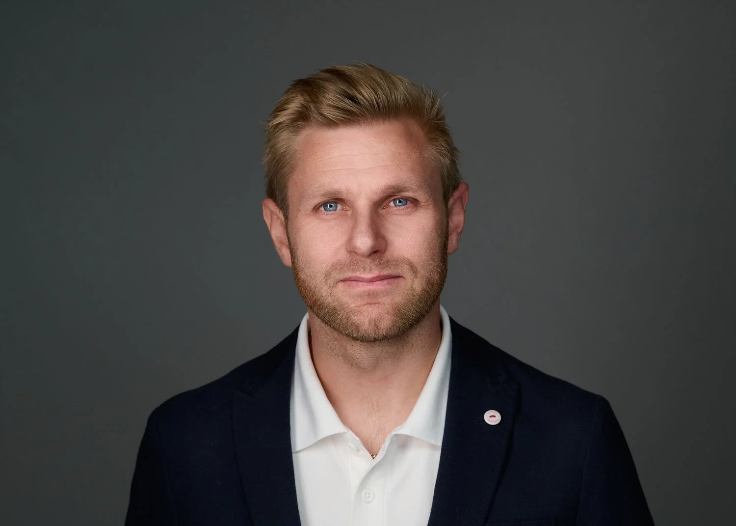 A headshot of a man with blonde hair and a beard, wearing a navy blazer and white shirt, against a gray background.