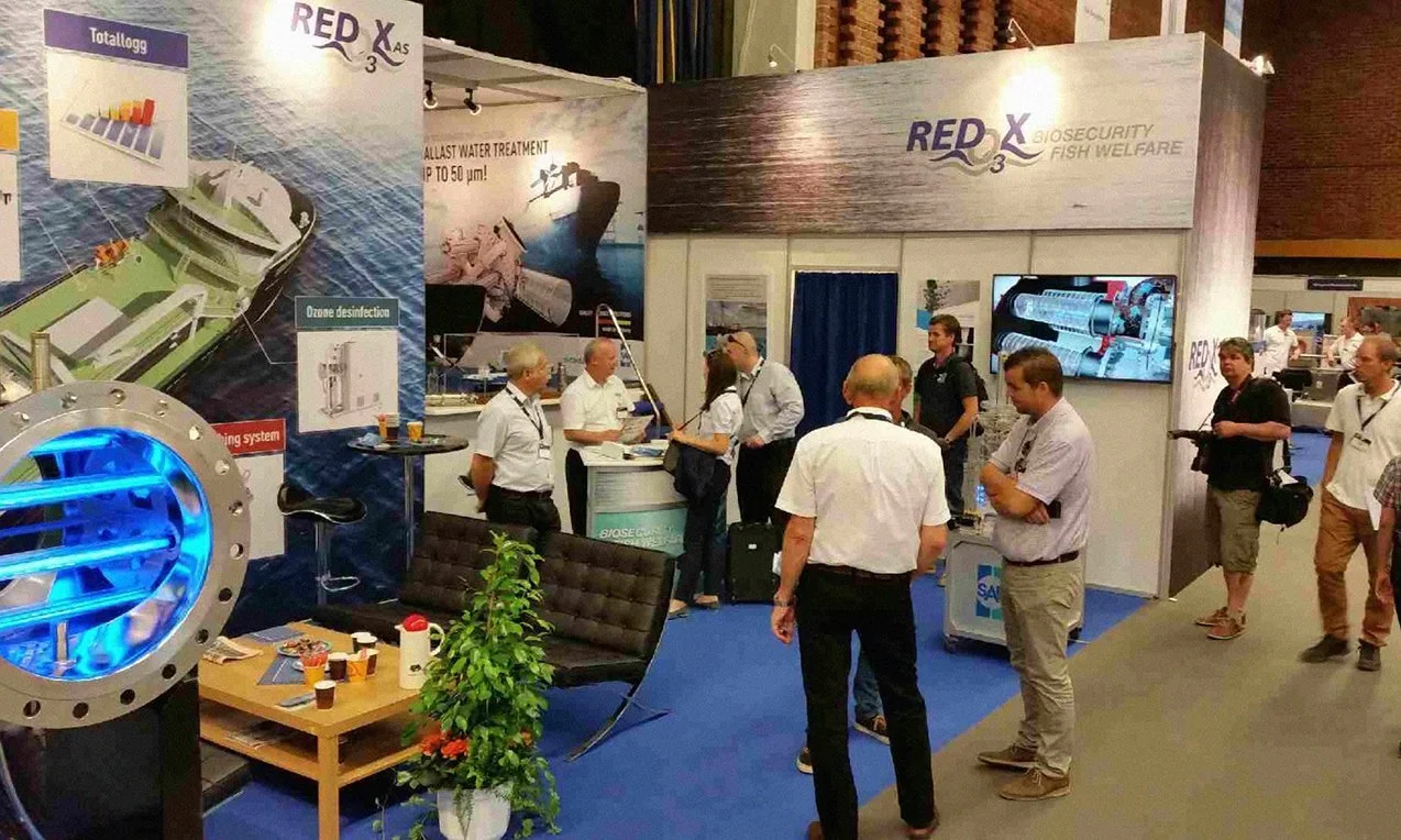 Trade show booth for RED X Biosecurity Fish Welfare featuring banners, informational displays, and a large tank with blue lighting, with attendees engaging and discussing.