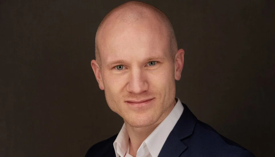 Portrett av en mann med blå øyne, lyst hud, kort hår og smil, iført en svart dress og hvit skjorte mot en mørk bakgrunn.