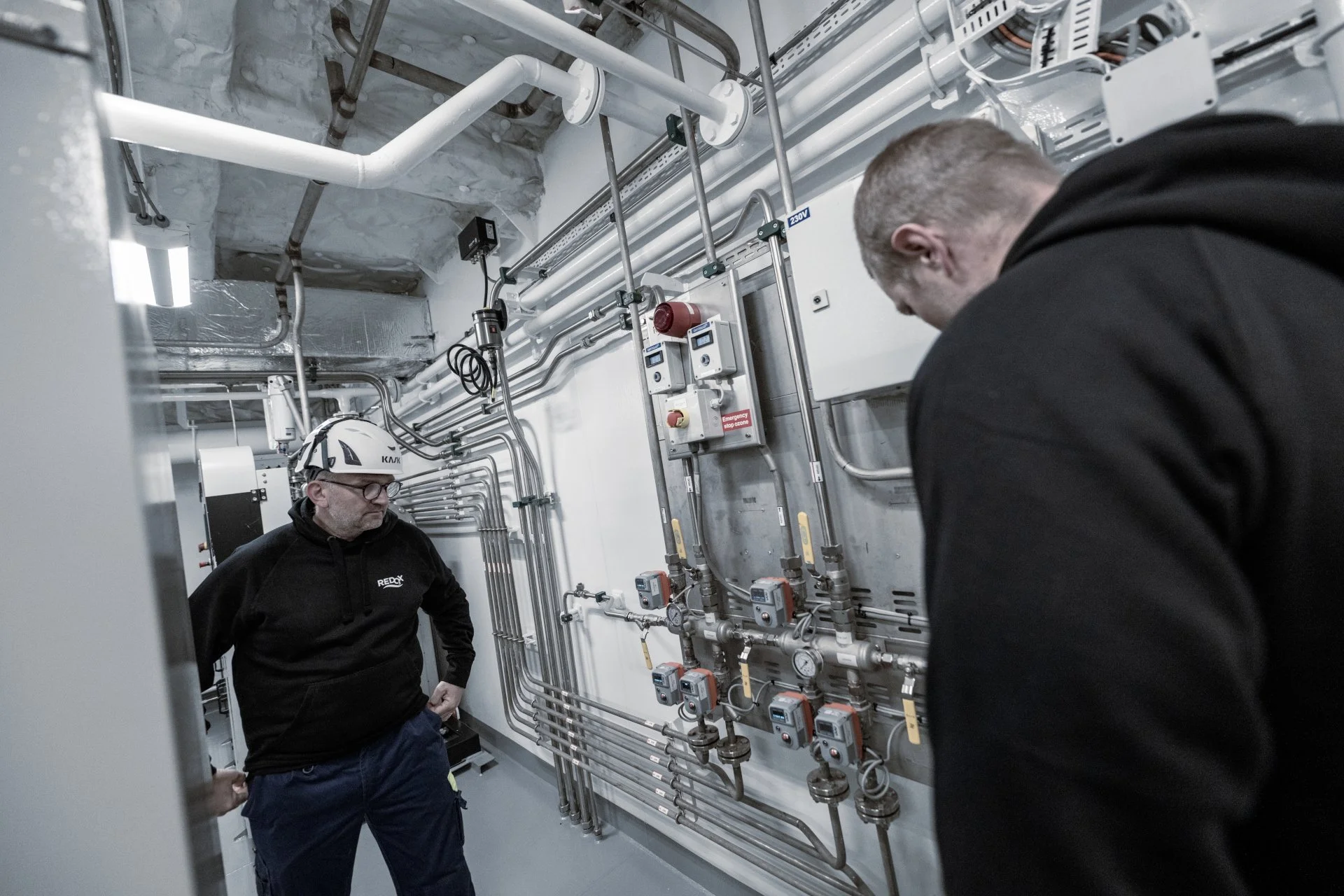 Two men examining a complex wall of pipes, gauges, and control panels in an industrial or mechanical room. One man is wearing a safety helmet with glasses, and the other has gray hair, both dressed in dark clothing.