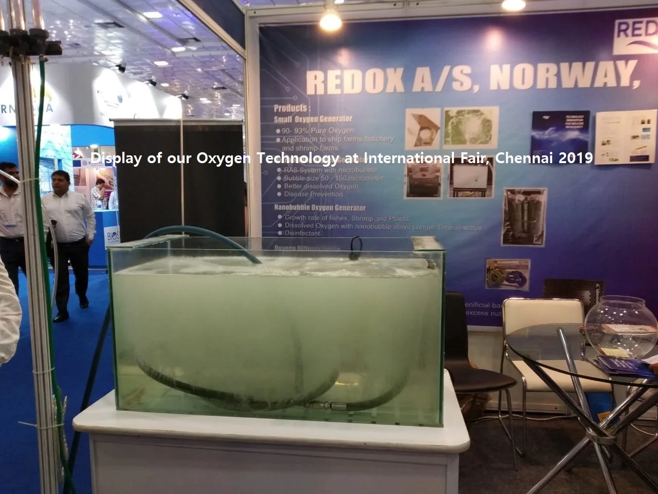 Utstilling av et lite oksygen-genereringsapparat på en messe, med en bakgrunnstavle som viser informasjon om produktet fra REDOX A/S, Norge. Flere mennesker går rundt i bakgrunnen.