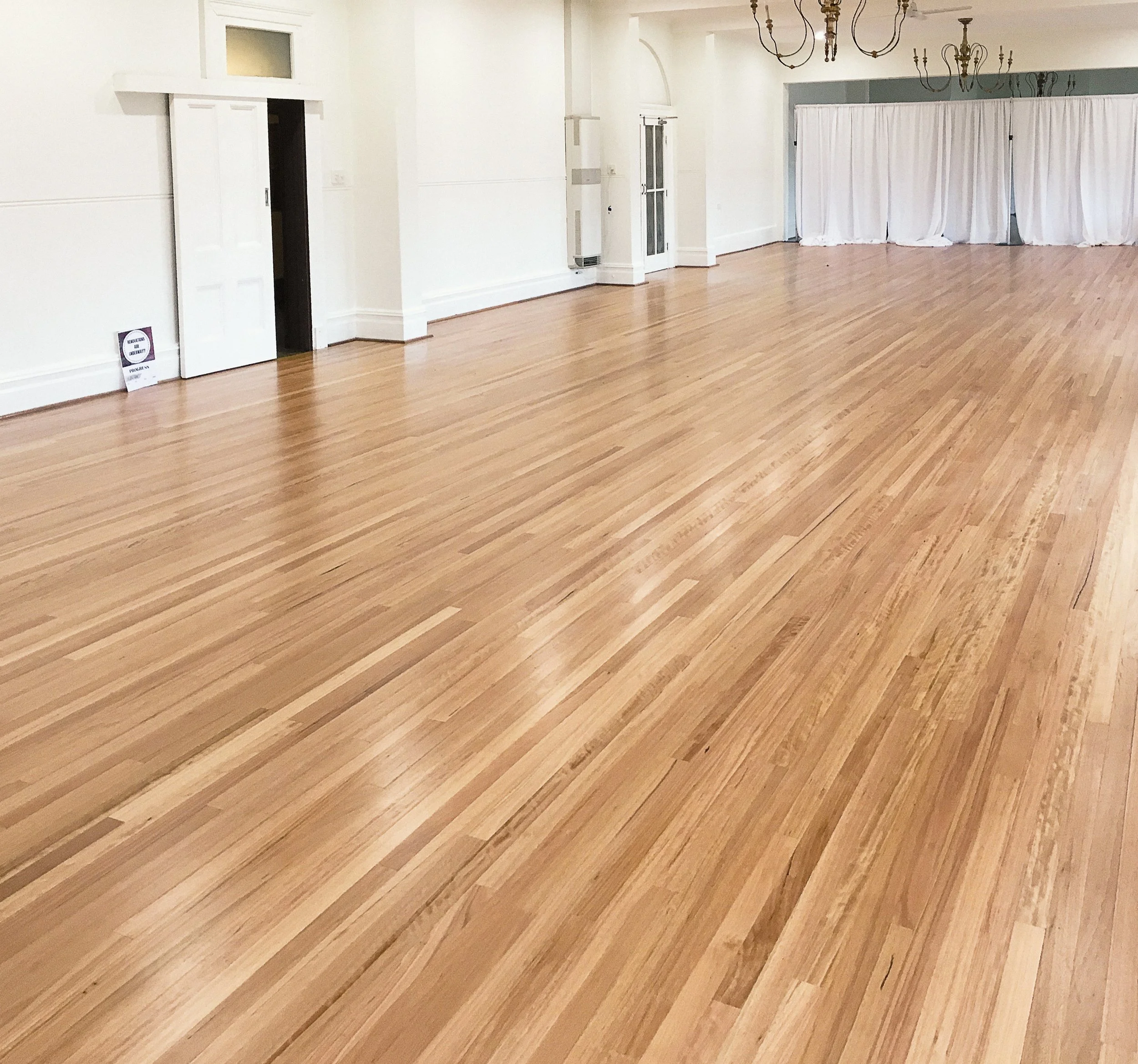 Spacious empty room with polished wooden floors, white walls, chandeliers, and a white curtain dividing part of the space.