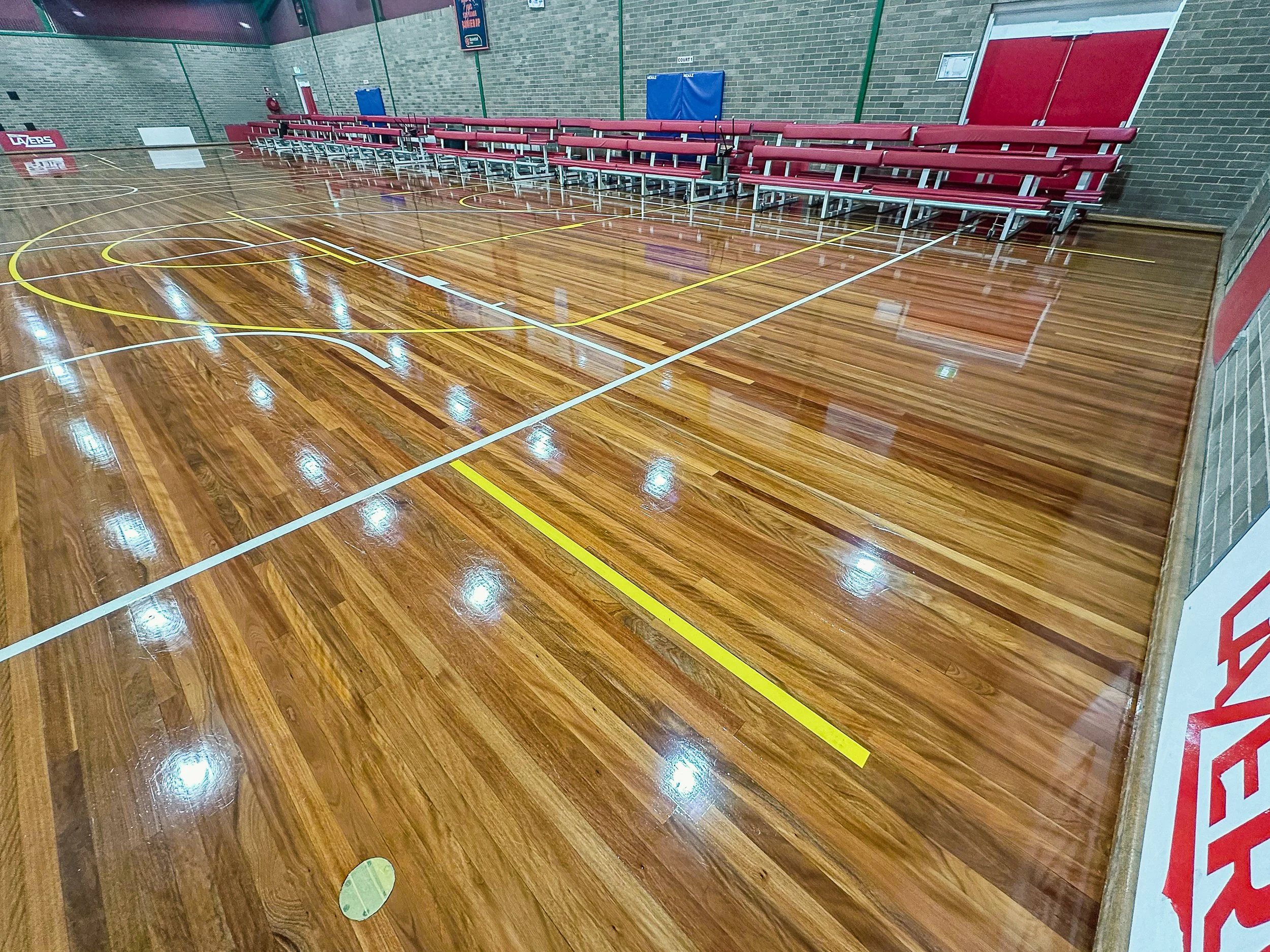Empty indoor basketball court with polished wooden flooring, red bleachers along the back wall, and a brick wall on the right side.