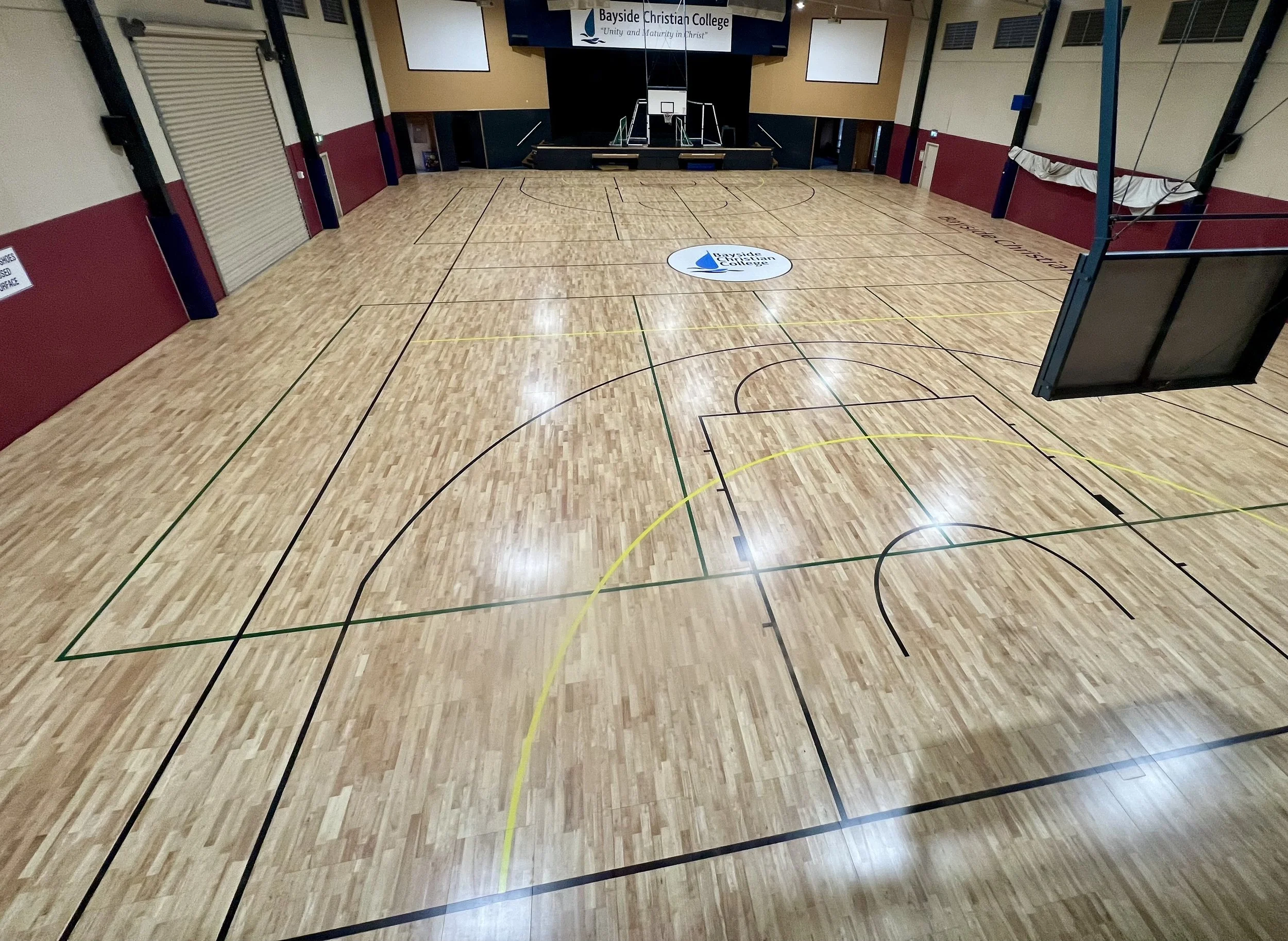 Empty indoor basketball court with a wooden floor, basketball hoop, and a Bayside Christian College sign on the floor.