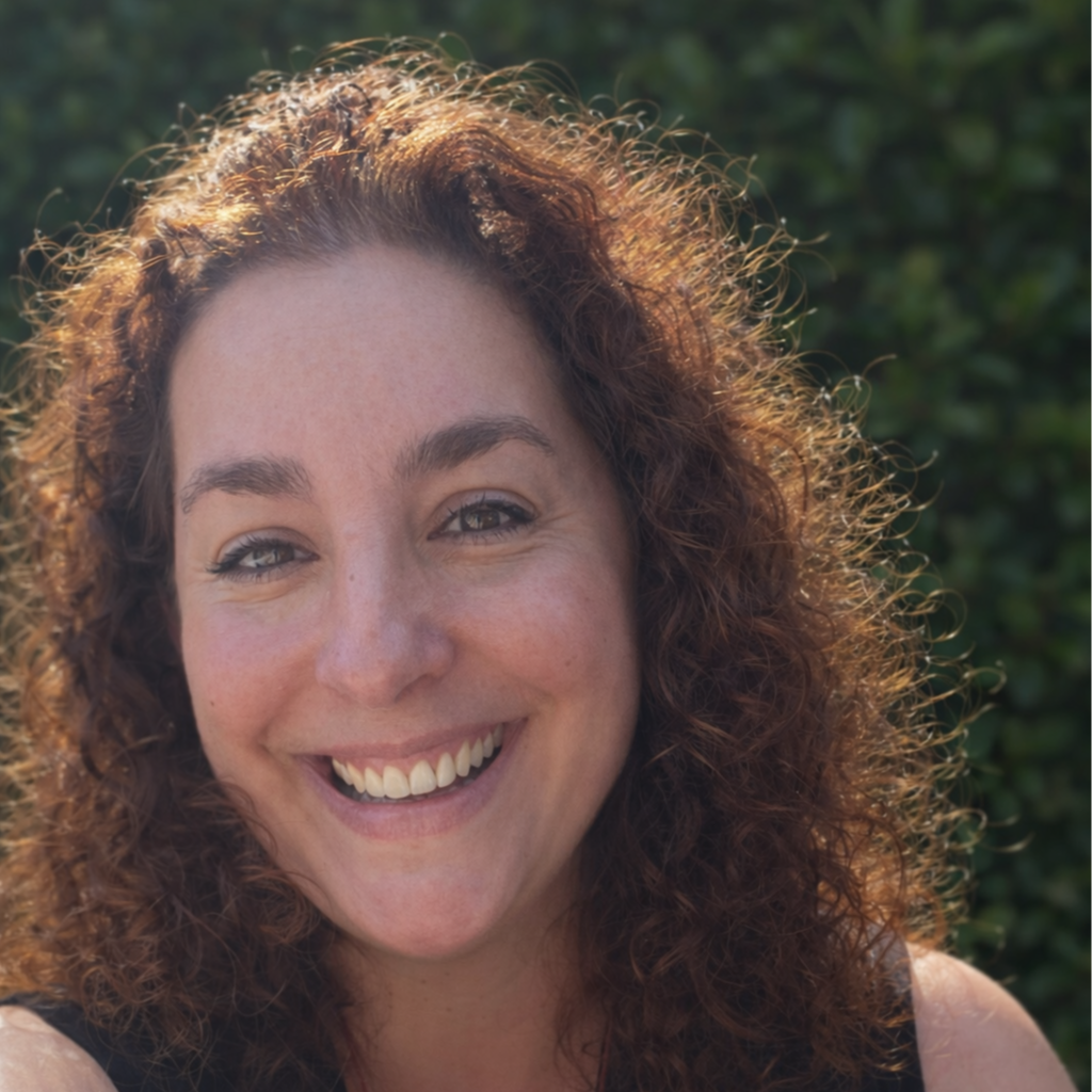 Femme souriante avec des cheveux roux, prise en extérieur avec un fond de végétation.