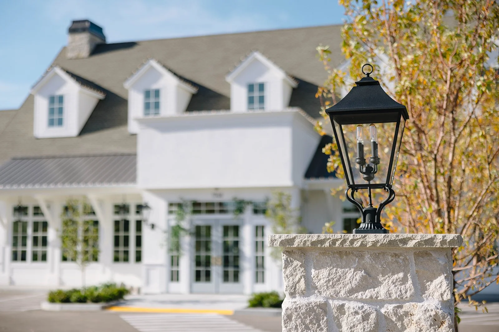 Close-up of a black metal lamp post in front of a white building with multiple windows and dormers, and a tree with autumn leaves.