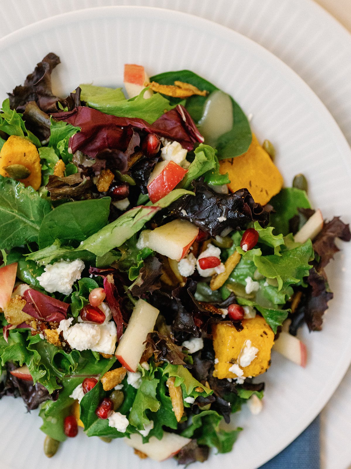 Mixed green salad with various vegetables, pomegranate seeds, sliced apples, crumbled cheese, and pumpkin seeds on a white plate.