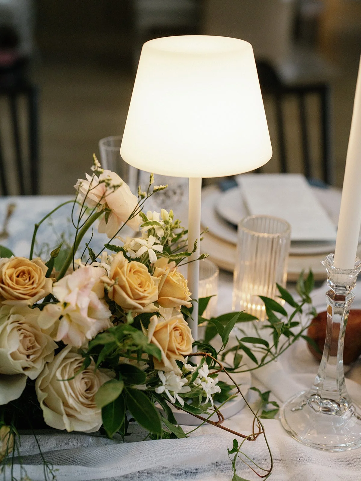 A table setting with a floral centerpiece, white roses, and greenery, illuminated by a white table lamp, with glassware and plates in the background.