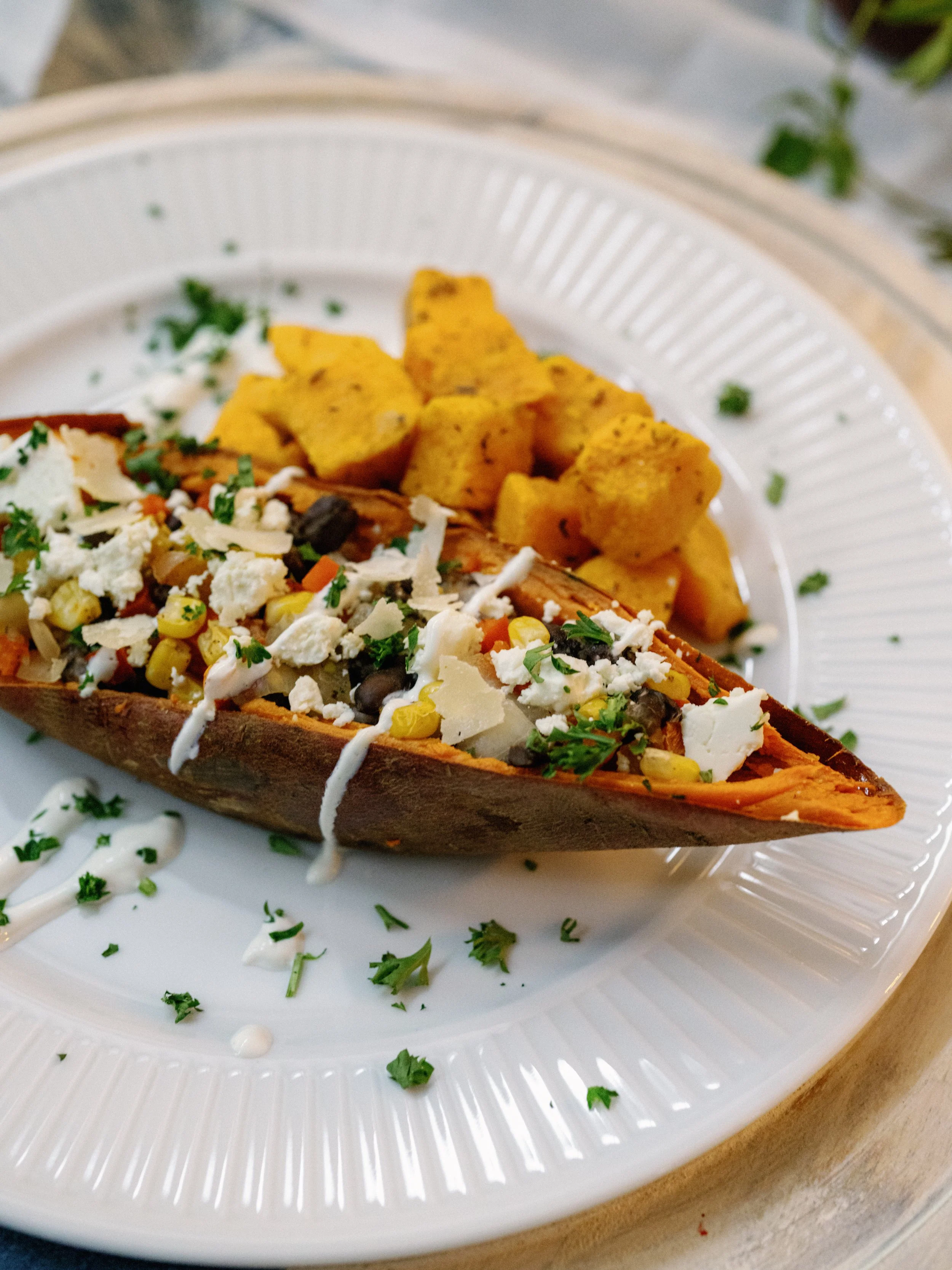 Sweet potato stuffed with black beans, corn, cheese, and chopped herbs, with a side of seasoned fried potatoes on a white plate.