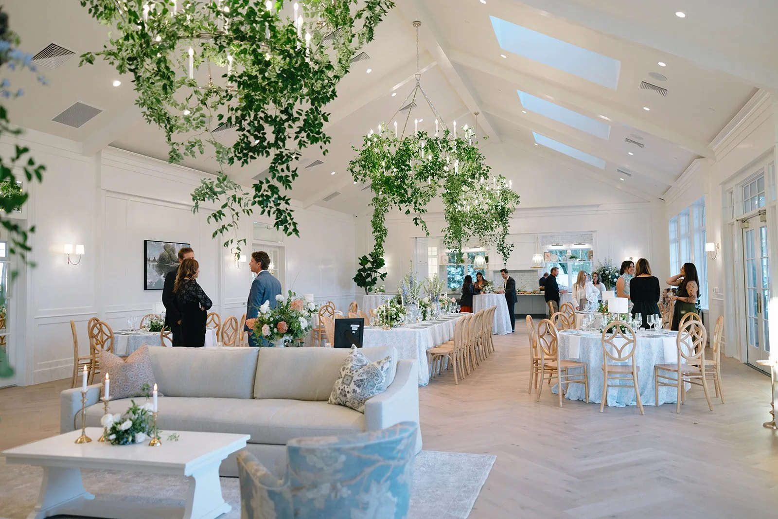 Elegant event space with white walls, large windows, and a vaulted ceiling adorned with hanging greenery and chandeliers. Multiple round tables with white tablecloths and wooden chairs are set for a meal. Guests are mingling and engaging in conversat